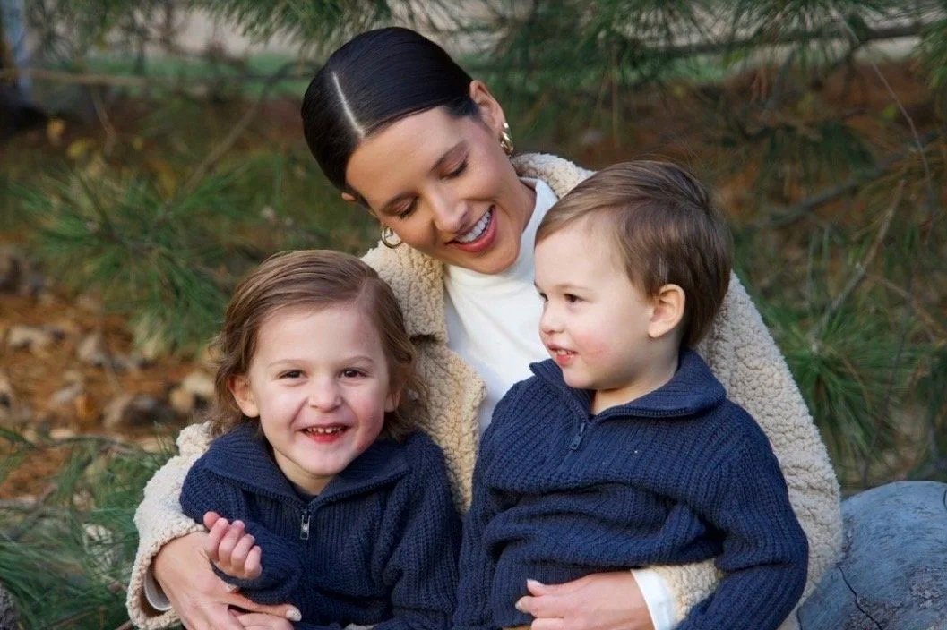A woman with two young children sitting outdoors in a wooded area, smiling and enjoying each other's company.