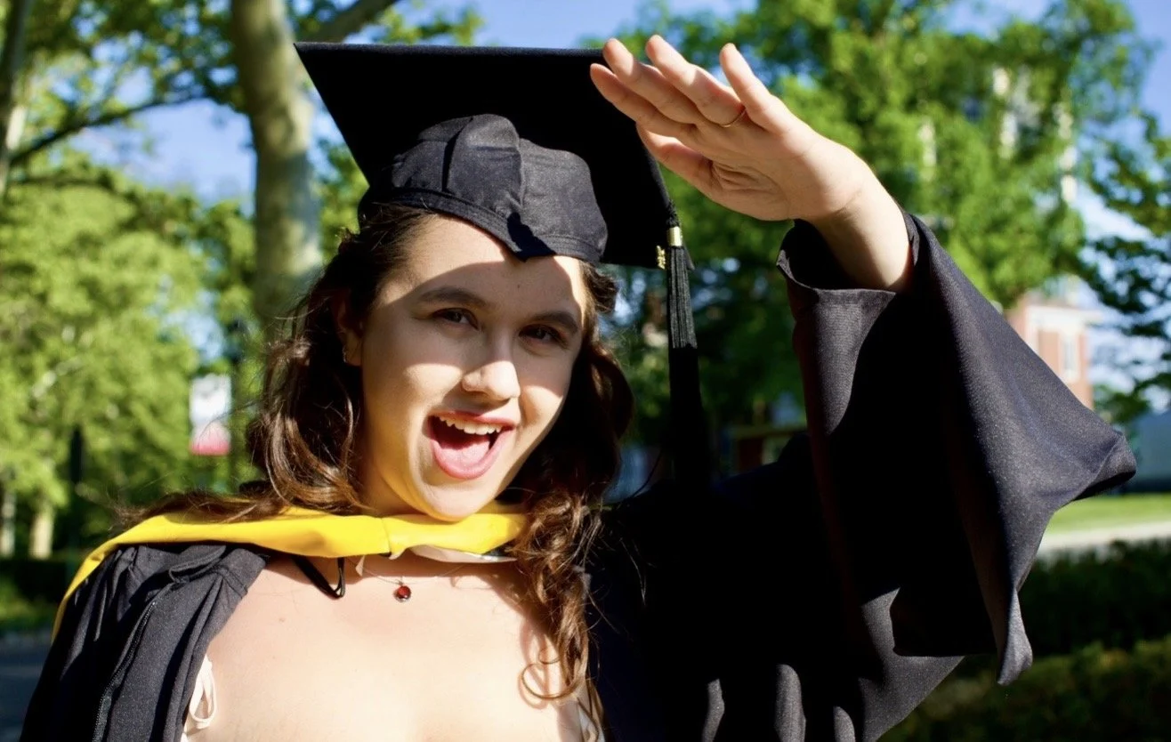 A young woman in graduation cap and gown celebrating outdoors, smiling and saluting with her right hand.