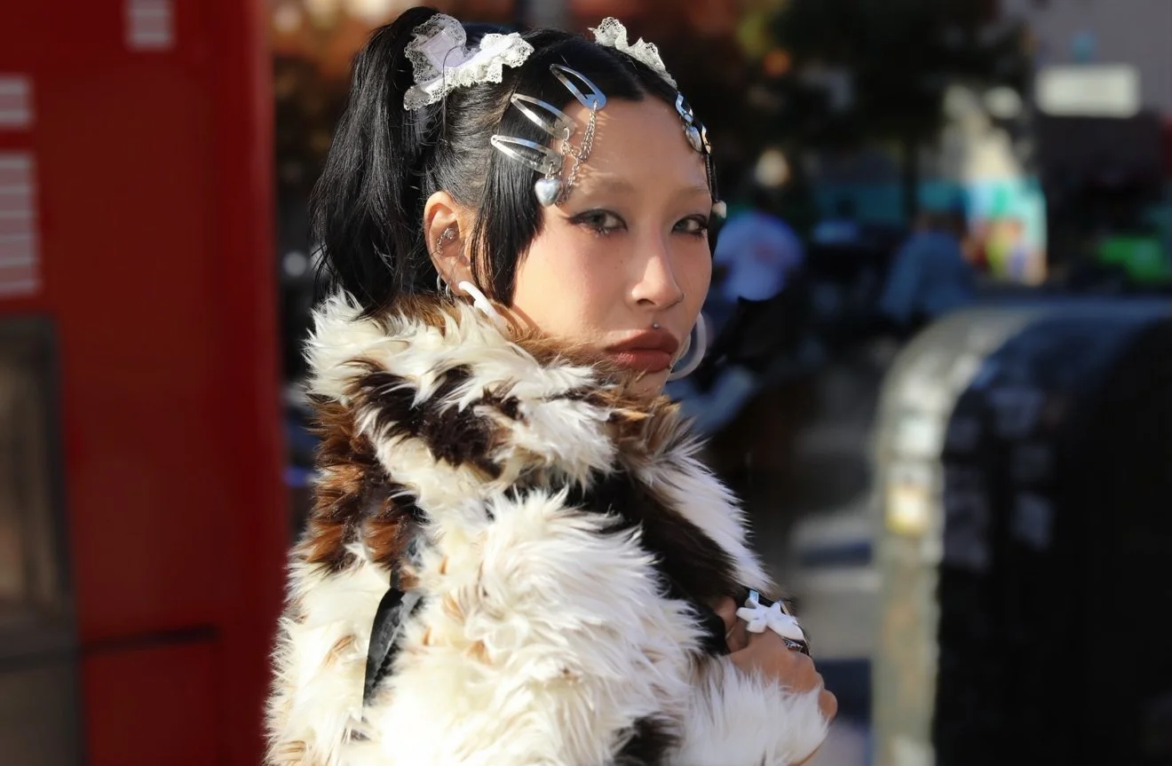 Young woman with dark hair adorned with hair clips and lace hair accessories, wearing a fur coat with a pattern of brown, black, and white, looking over her shoulder on a city street.