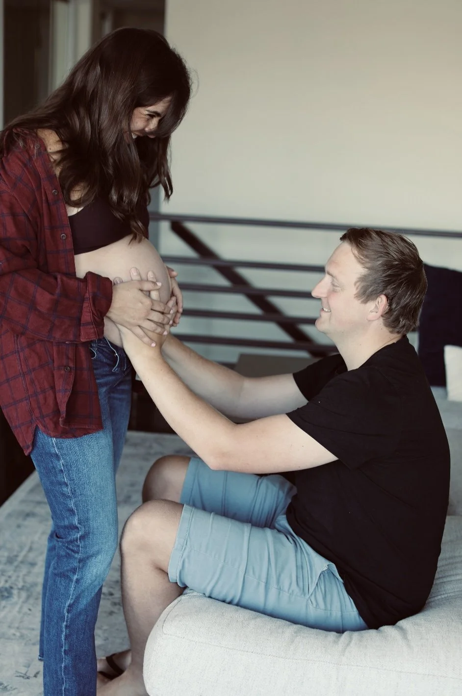 Pregnant woman standing in front of man sitting on a chair, with both holding her belly and smiling.