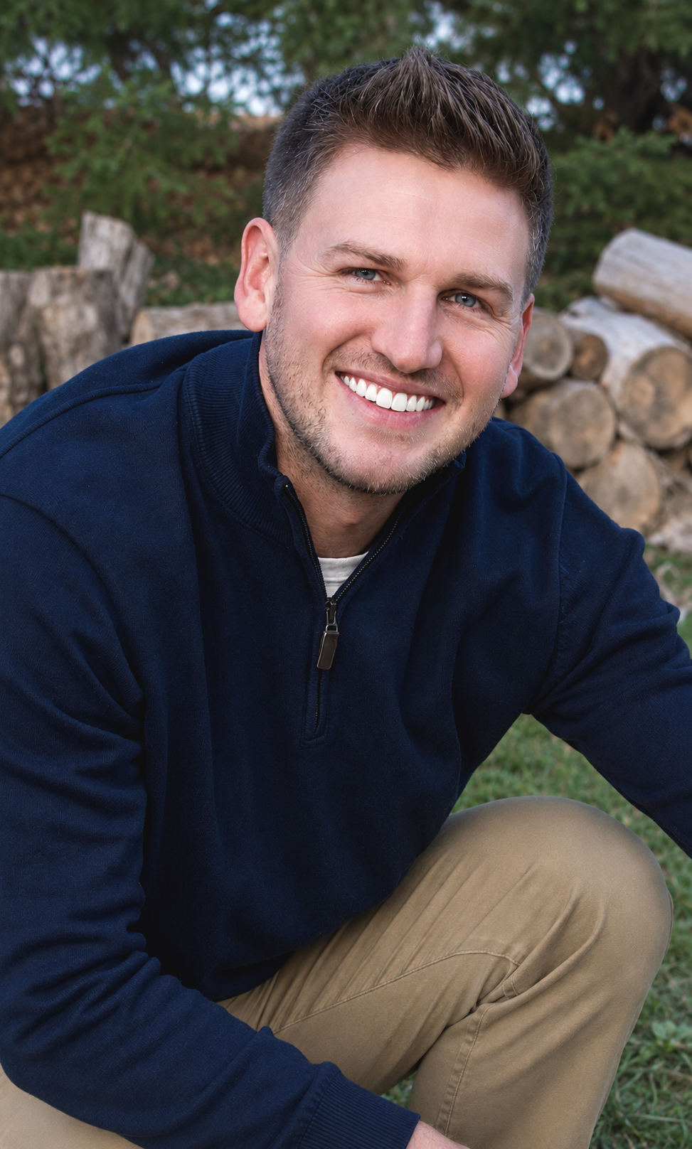 A young man with short brown hair and blue eyes smiling outdoors, sitting on grass with stacked logs and greenery in the background.