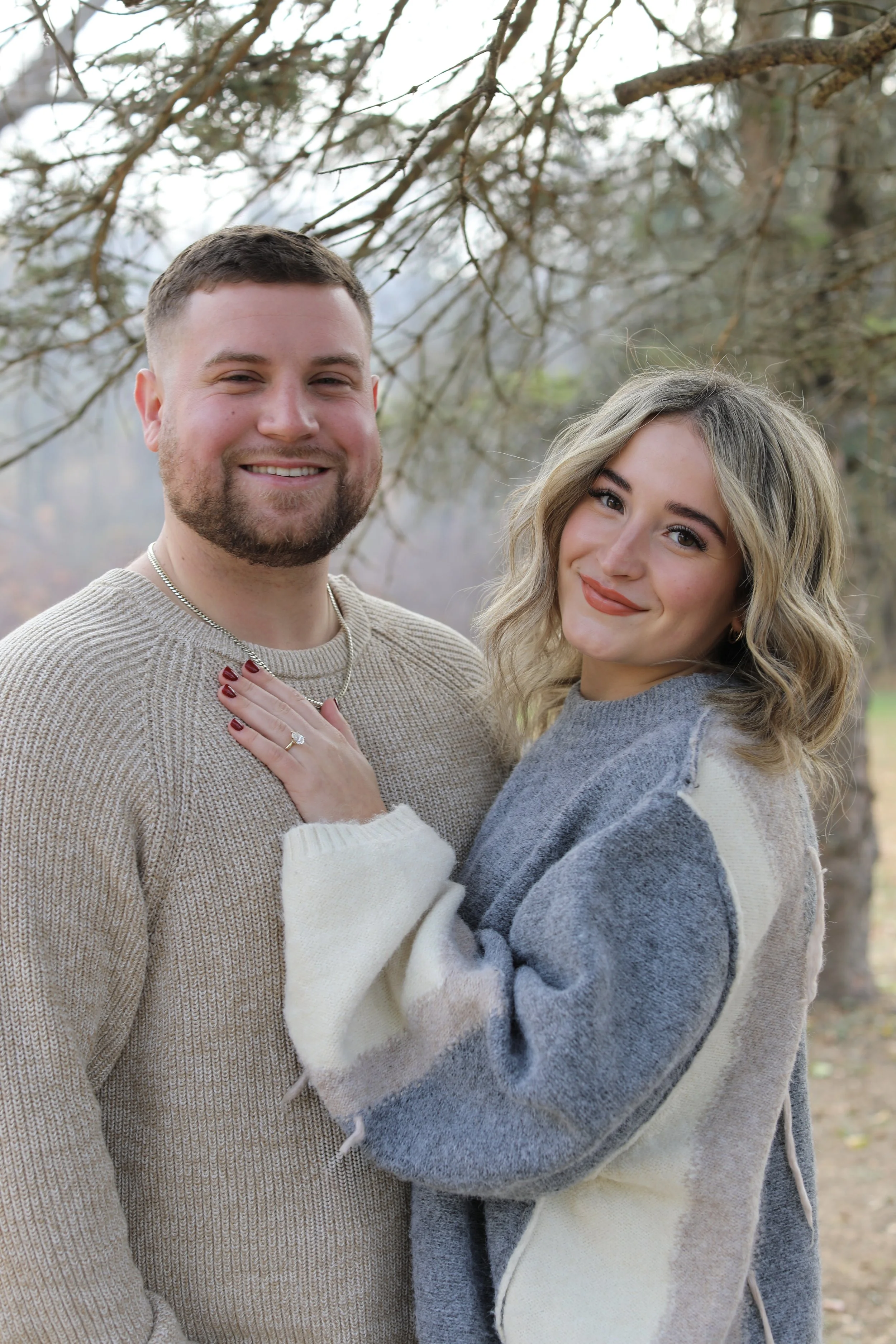 A smiling man and woman standing outdoors among bare trees, dressed in warm sweaters, embracing each other.
