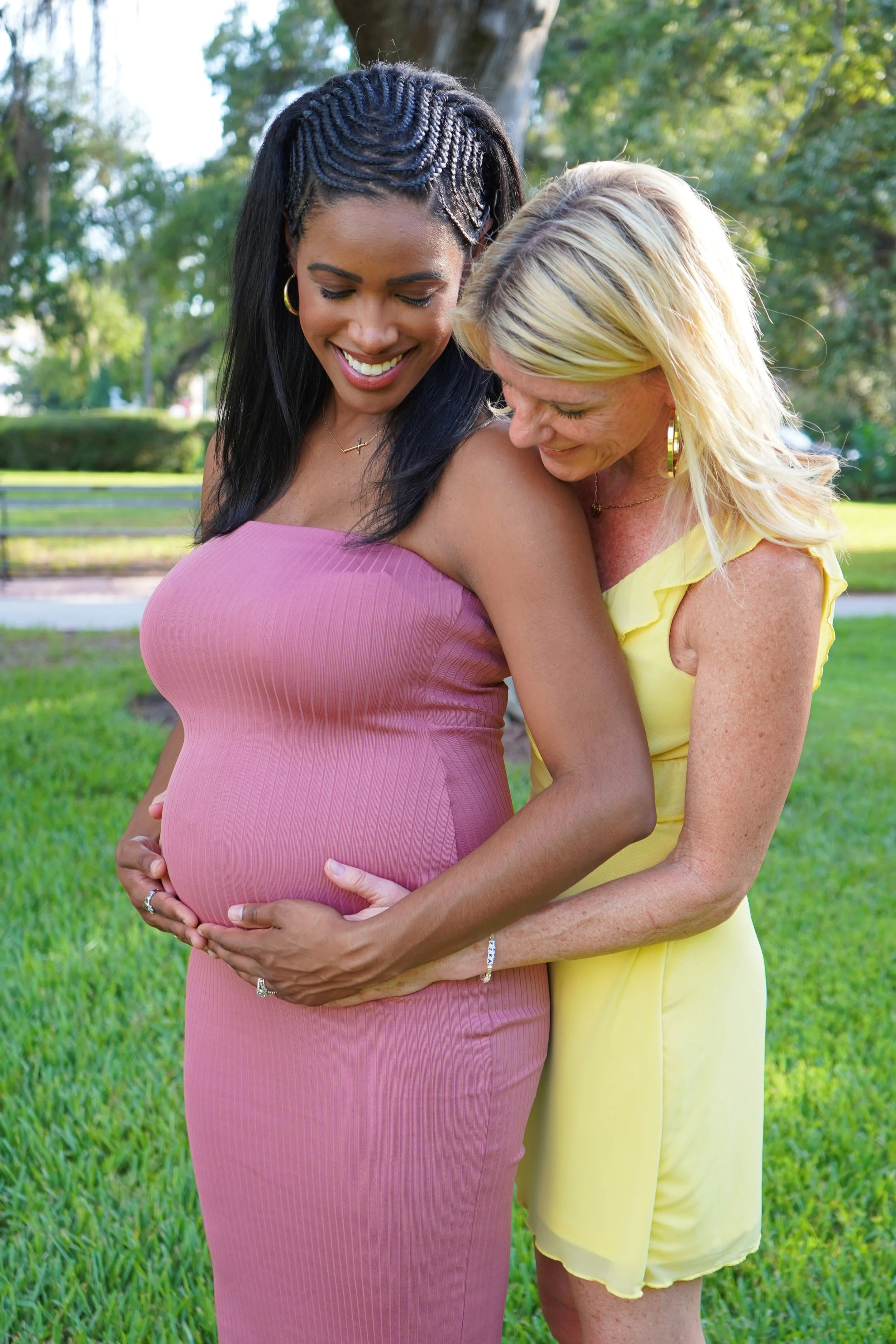 A pregnant woman in a pink dress and a woman in a yellow dress are embracing outdoors in a park with green grass and trees, with the pregnant woman holding her belly and both smiling.