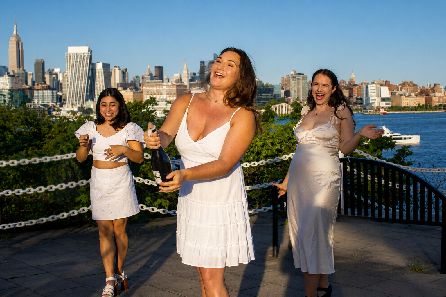 Three women celebrating outdoors, one opening a bottle of champagne, with a city skyline and river in the background.