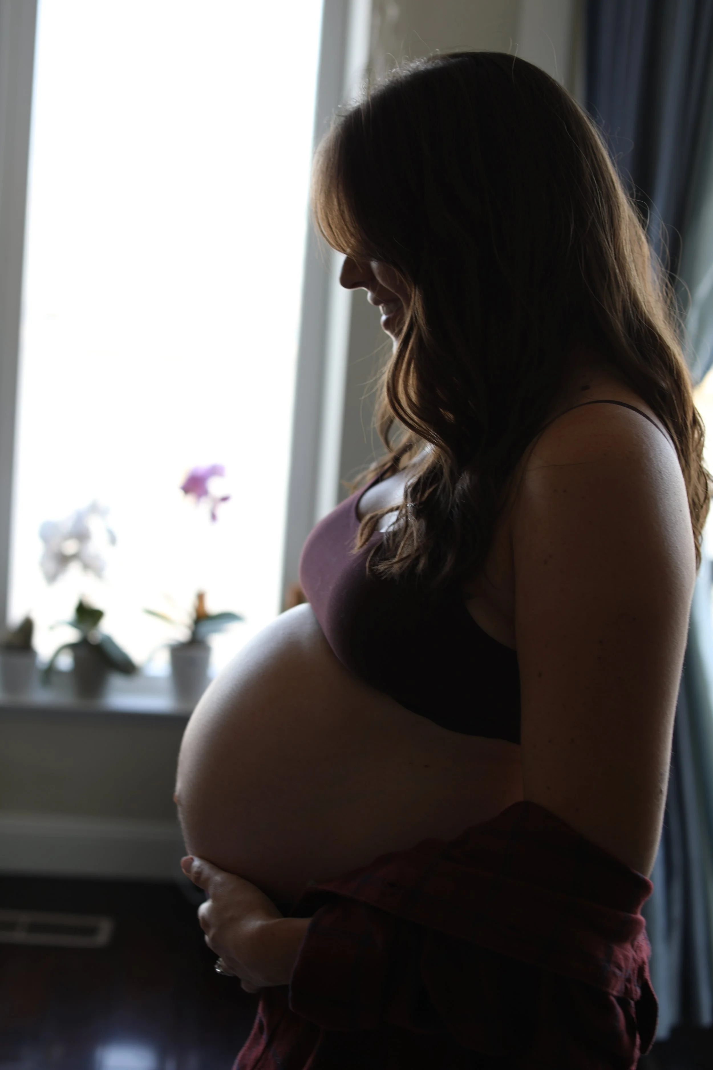 A pregnant woman standing indoors, smiling and looking down, with natural light coming from a window behind her, and houseplants on a windowsill in the background.
