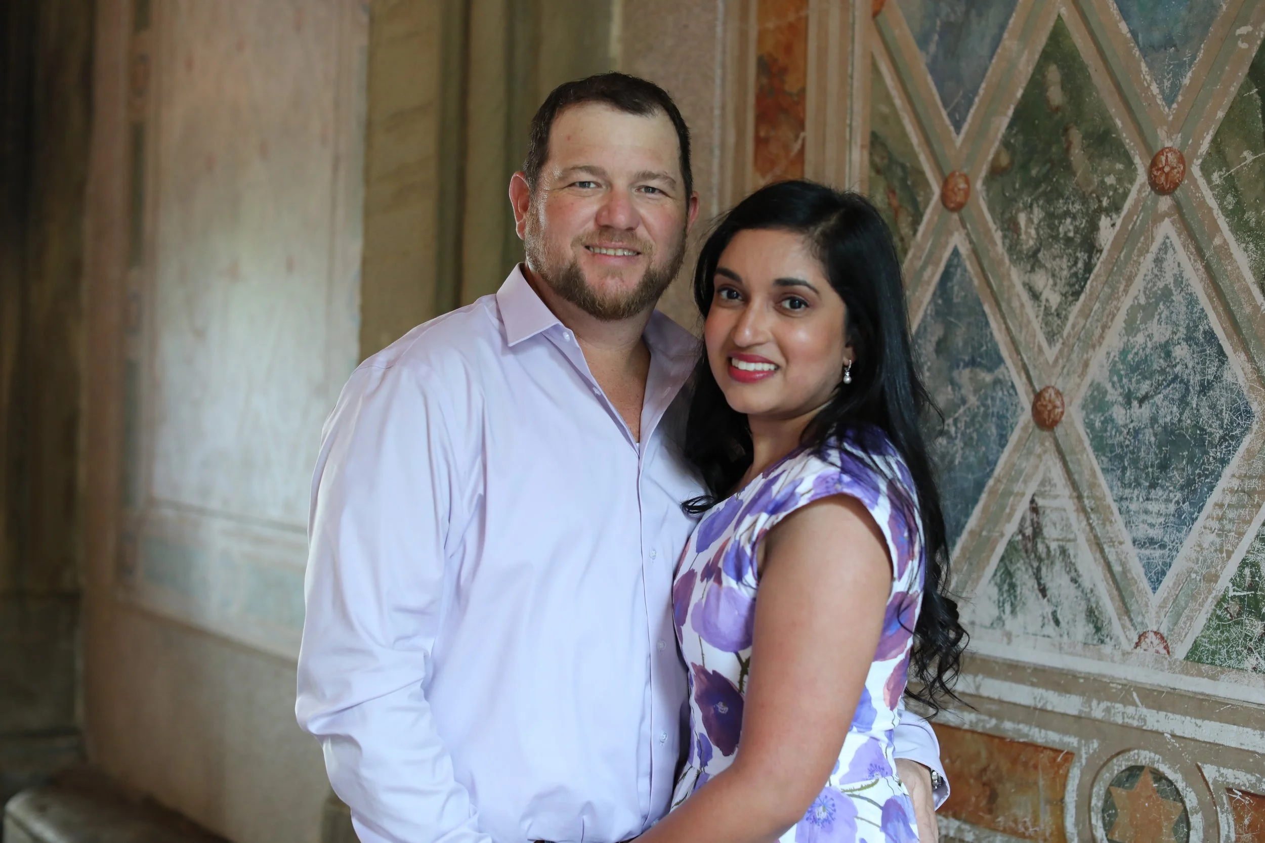 A smiling man and woman posing together against a decorative wall with geometric and floral patterns.