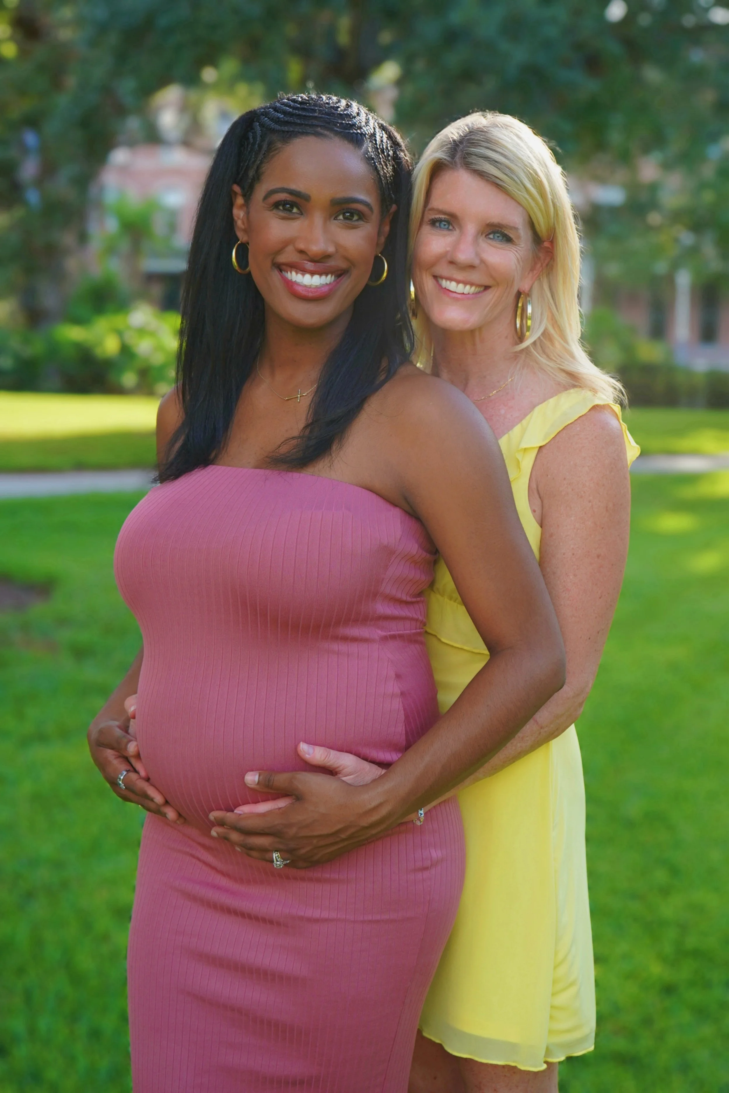 Two women smiling outdoors, one pregnant, embracing each other, with trees and grass in the background.