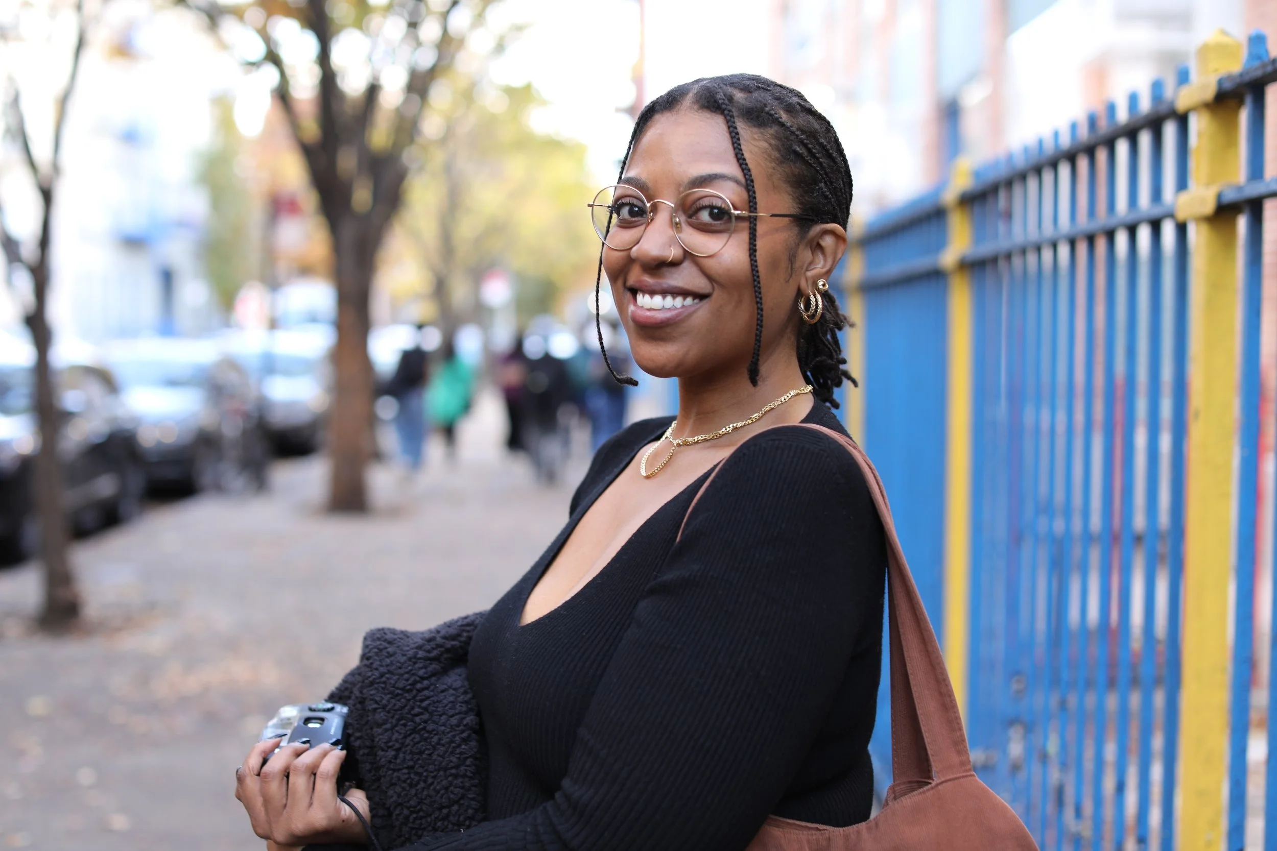 A smiling woman with glasses and earrings outdoors, holding a camera, with a colorful fence and trees in the background.