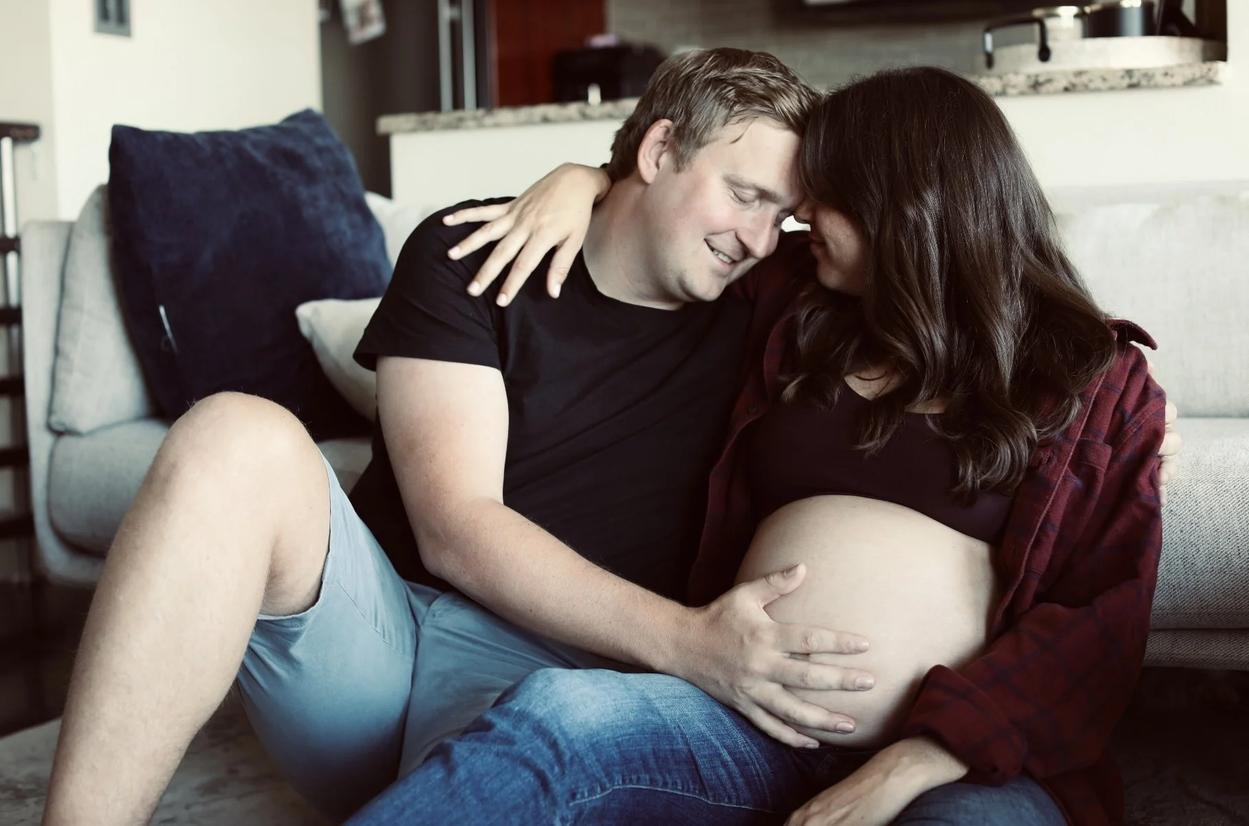 A couple sitting on a couch, tenderly touching and embracing an expecting woman with a pregnant belly, smiling with their foreheads touching in a cozy living room.