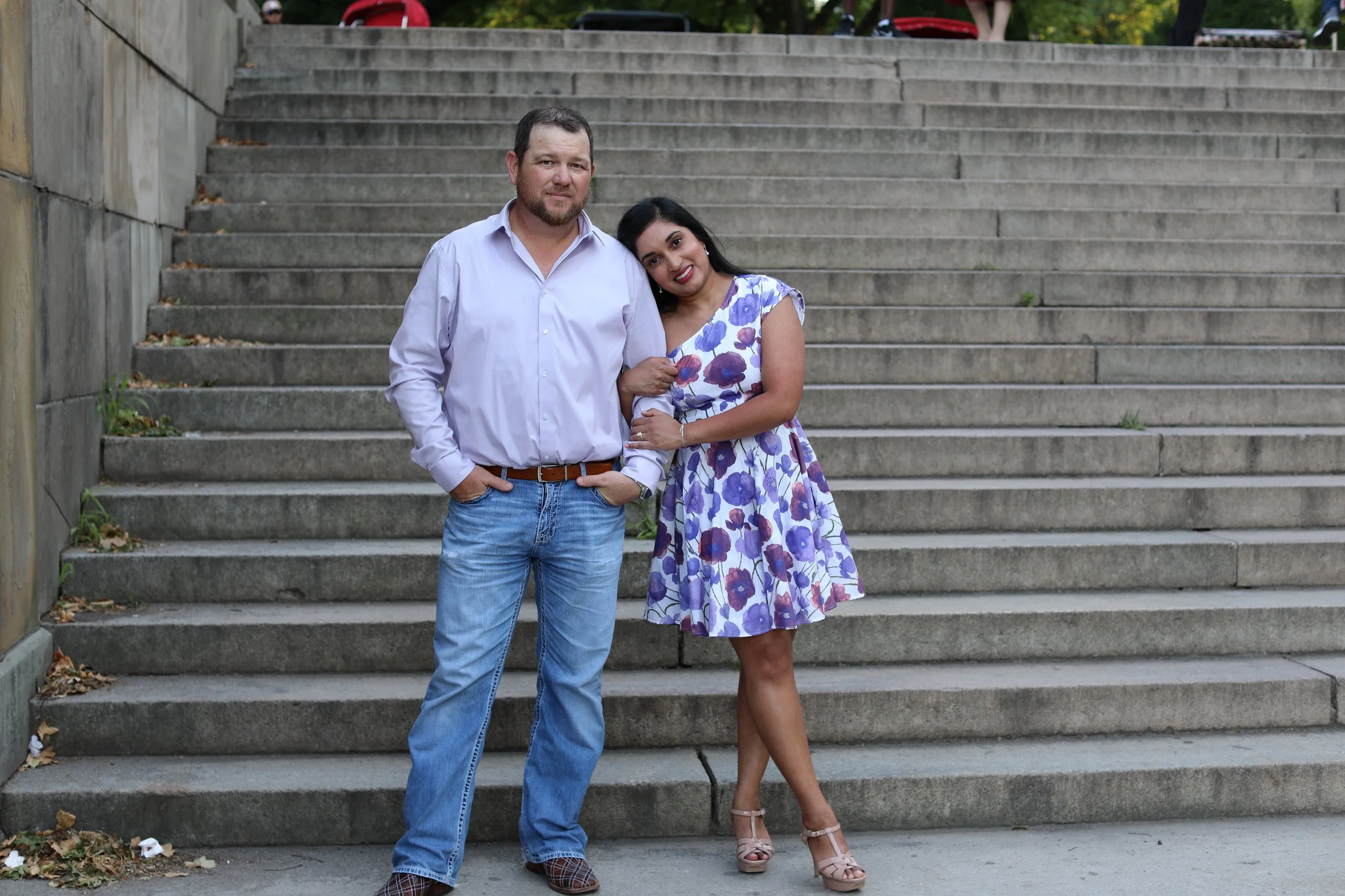 A man and woman standing arm-in-arm on outdoor concrete stairs. The man is wearing a light purple shirt and jeans, while the woman is in a floral dress with purple and pink flowers. They are smiling; the woman is leaning her head on the man's shoulde