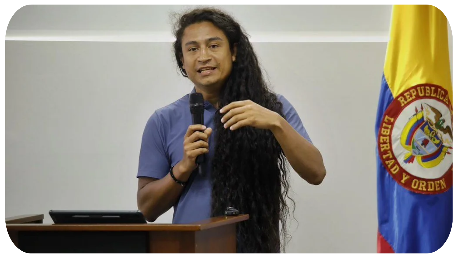 A man with long, curly black hair and medium skin tone speaking into a microphone at a podium, with a Venezuela flag partially visible on his right.