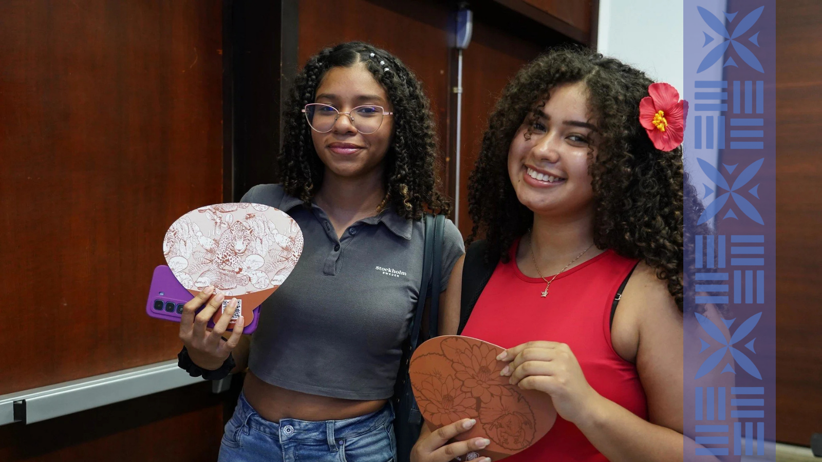 Two young women standing indoors, smiling. The woman on the left has curly hair, glasses, and is wearing a gray shirt with the word 'Stockholm' on it, holding a pillow with a paper background. The woman on the right has curly hair, a pink flower in her hair, and a red top, holding a pillow with a floral pattern.