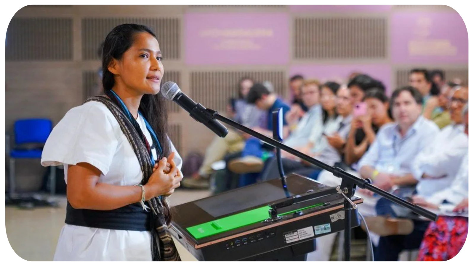 A woman speaking into a microphone at a podium during a conference or seminar, with an audience of diverse attendees listening attentively.