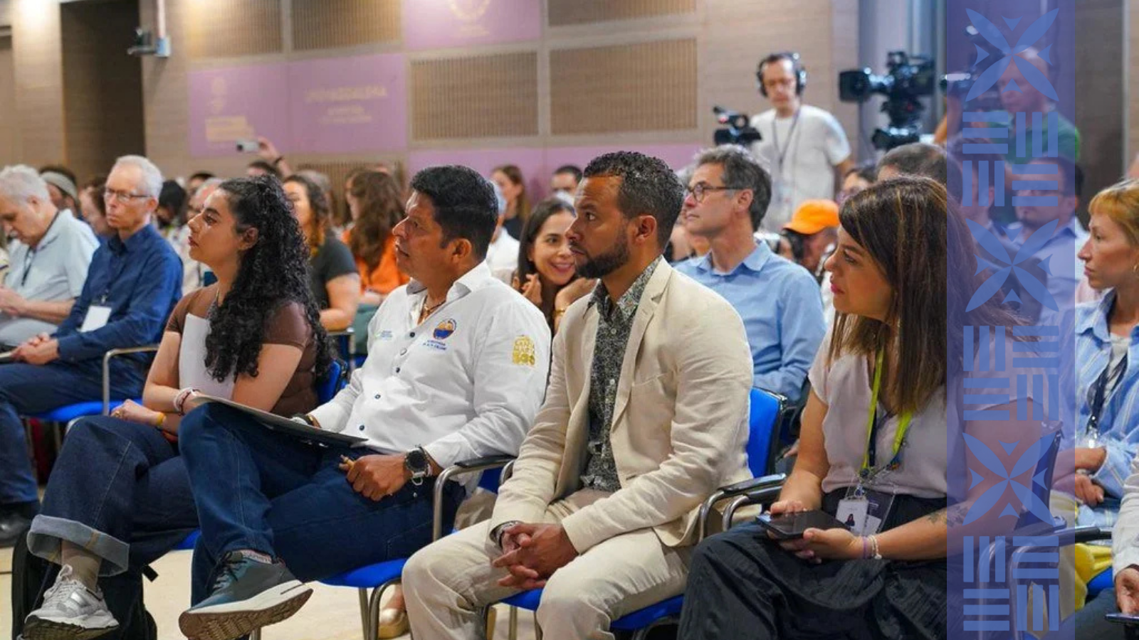 People attending a conference sitting in chairs, including a man in a cream suit, a woman with long dark hair in a white shirt, a man in a white shirt with a logo, and a woman with glasses and a notepad, with cameras and a speaker in the background.
