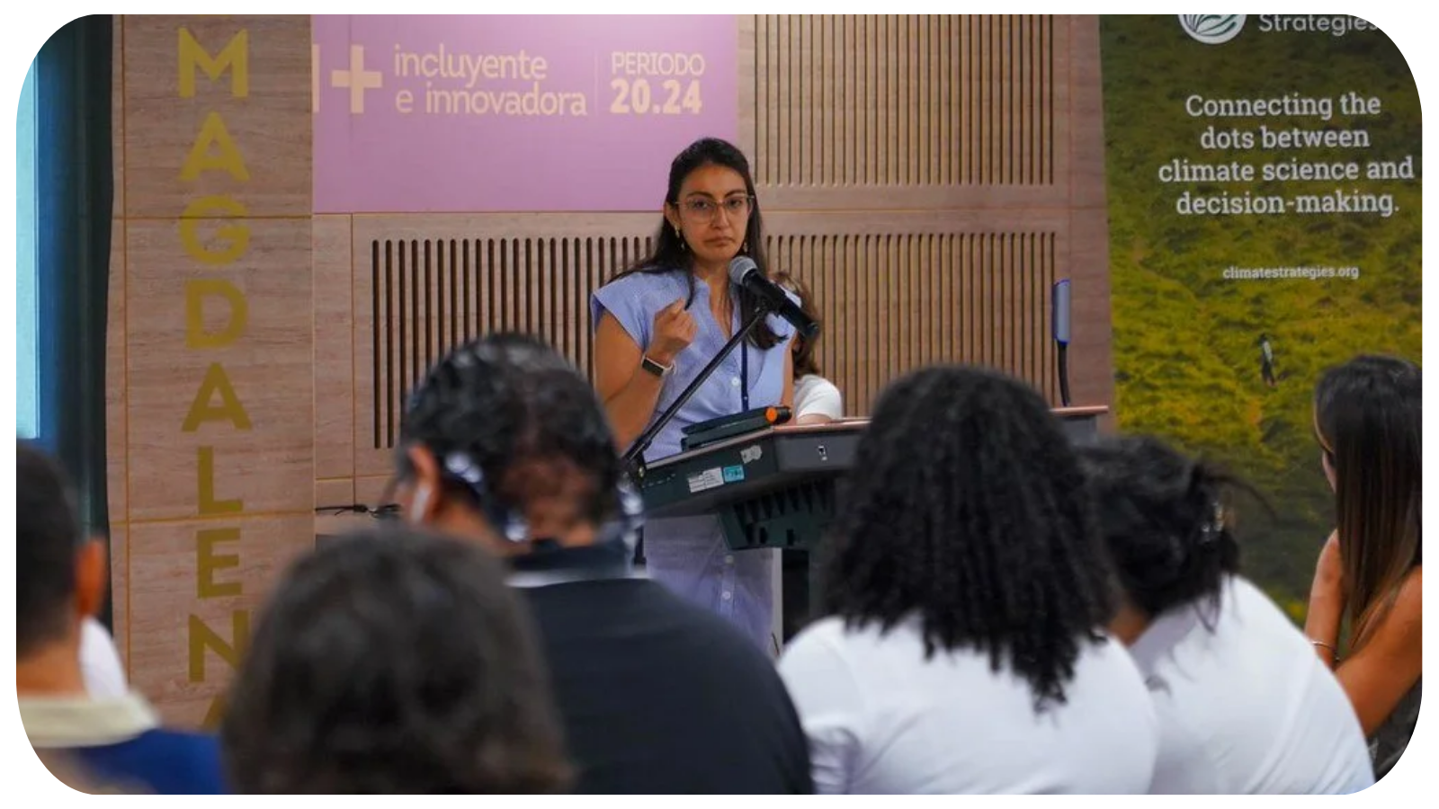 A woman standing at a podium speaking into a microphone during a conference with an audience. Behind her, banners display texts about climate strategies and inclusivity.