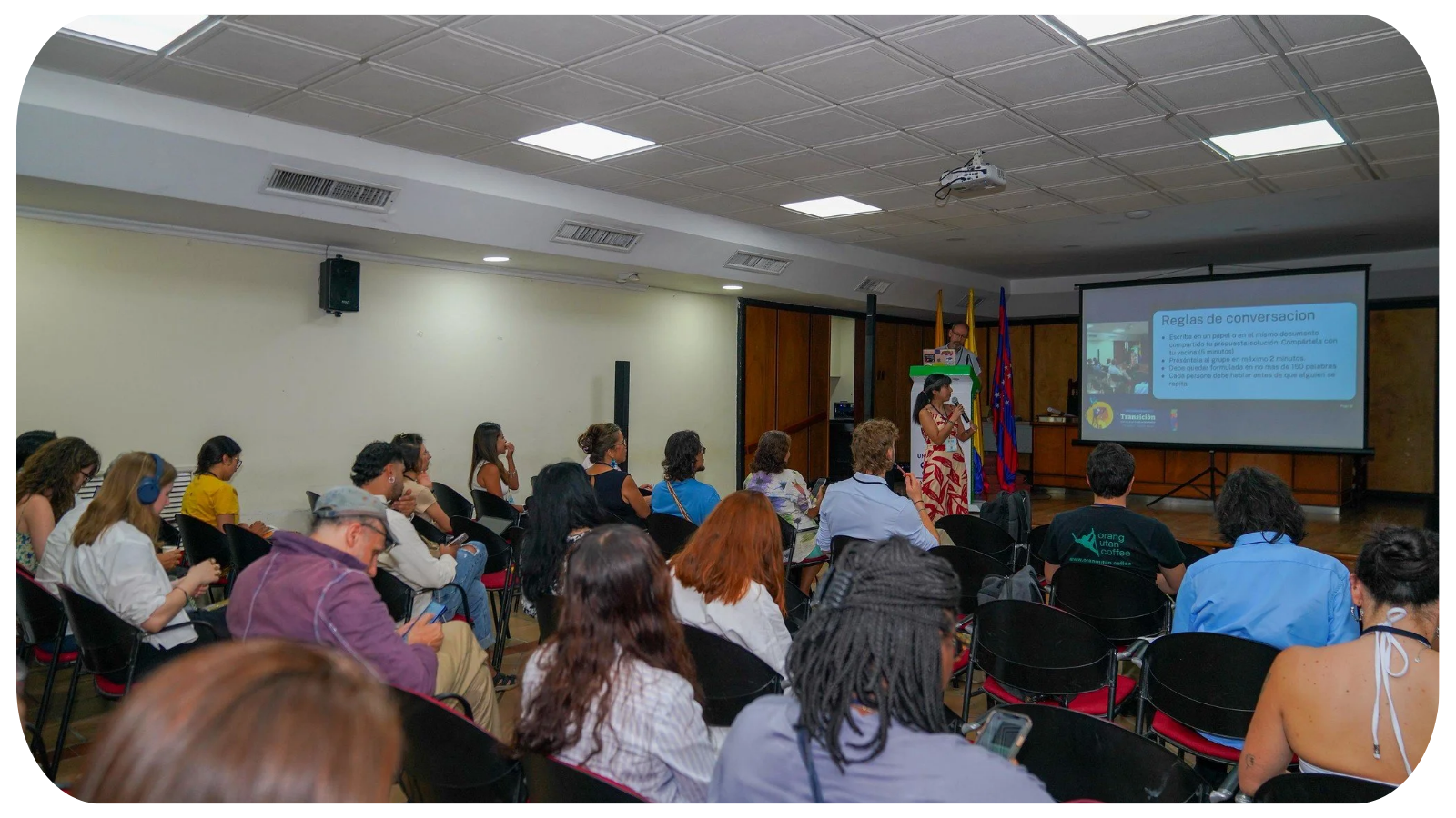 People attending a presentation in a conference room, with a woman speaking at the front and a large screen displaying presentation slides.