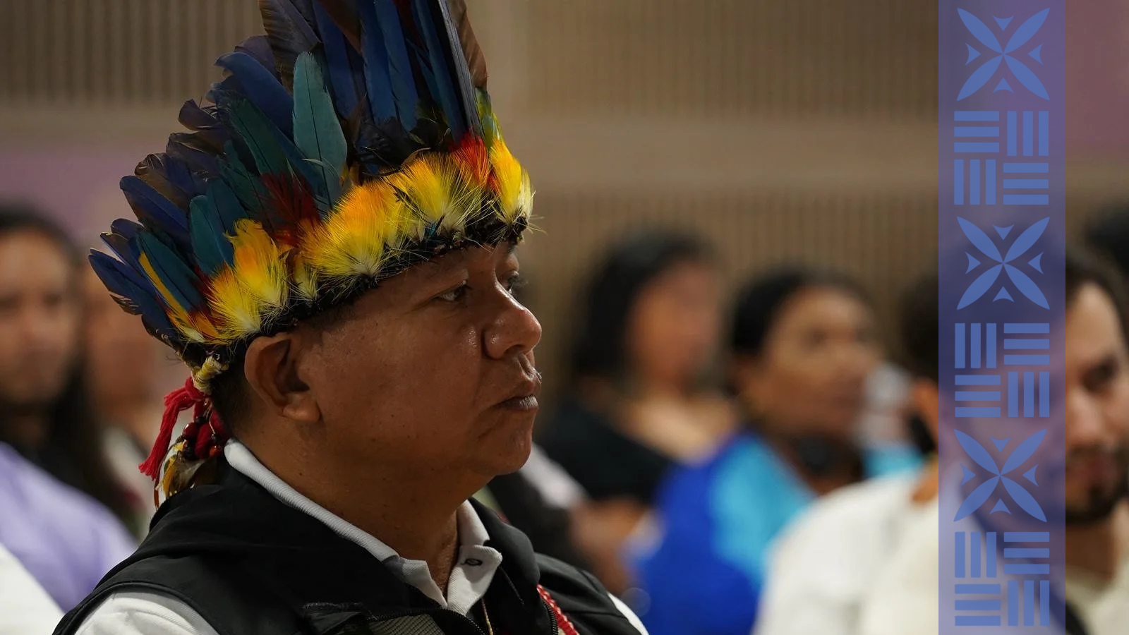 A man wearing a colorful feathered headdress participating in an indoor event, with a blurred audience in the background.