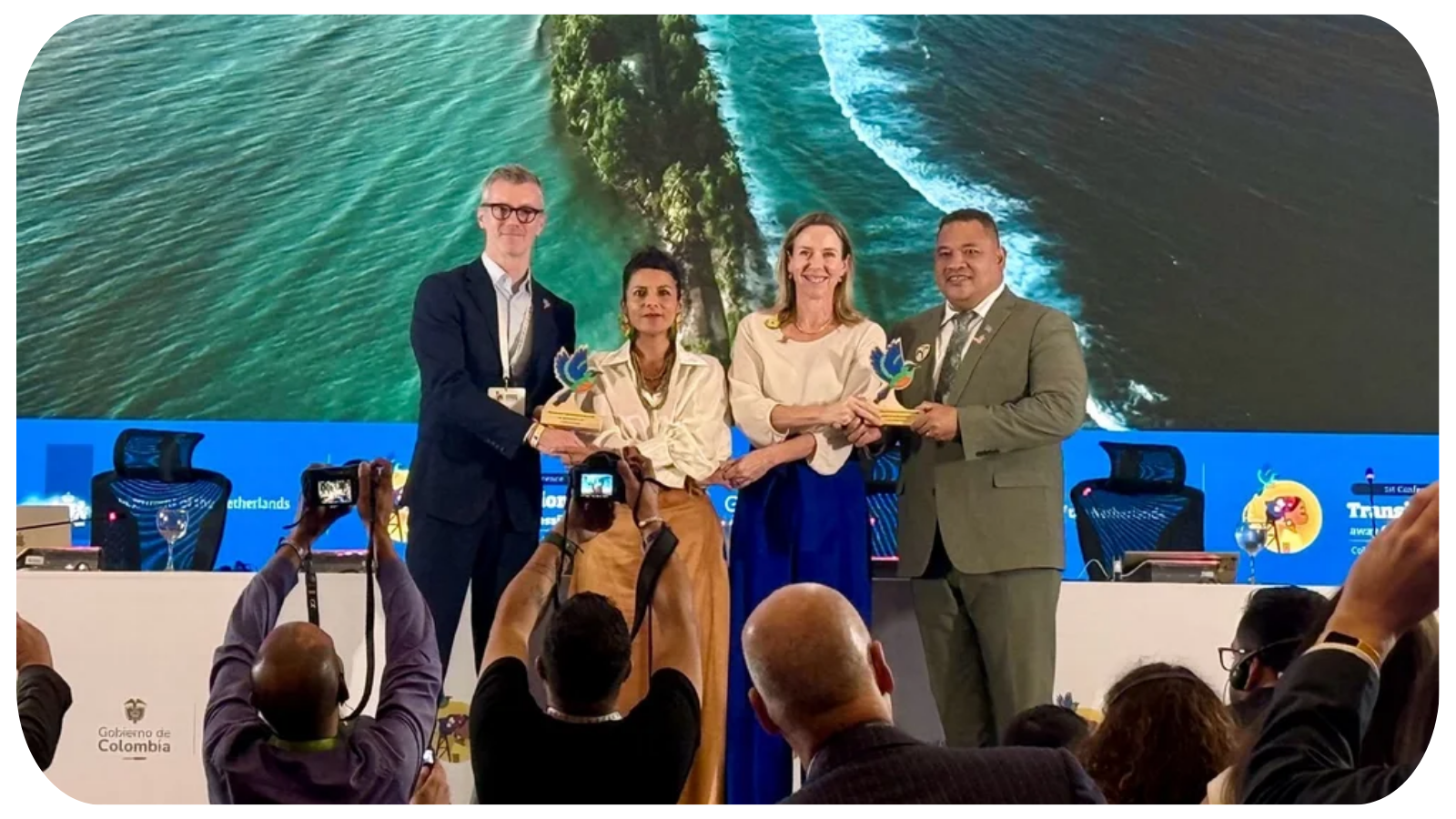 Four people on stage during an award ceremony, holding trophies with a large screen behind them displaying an aerial view of a coastline and water. Audience members are taking photos.
