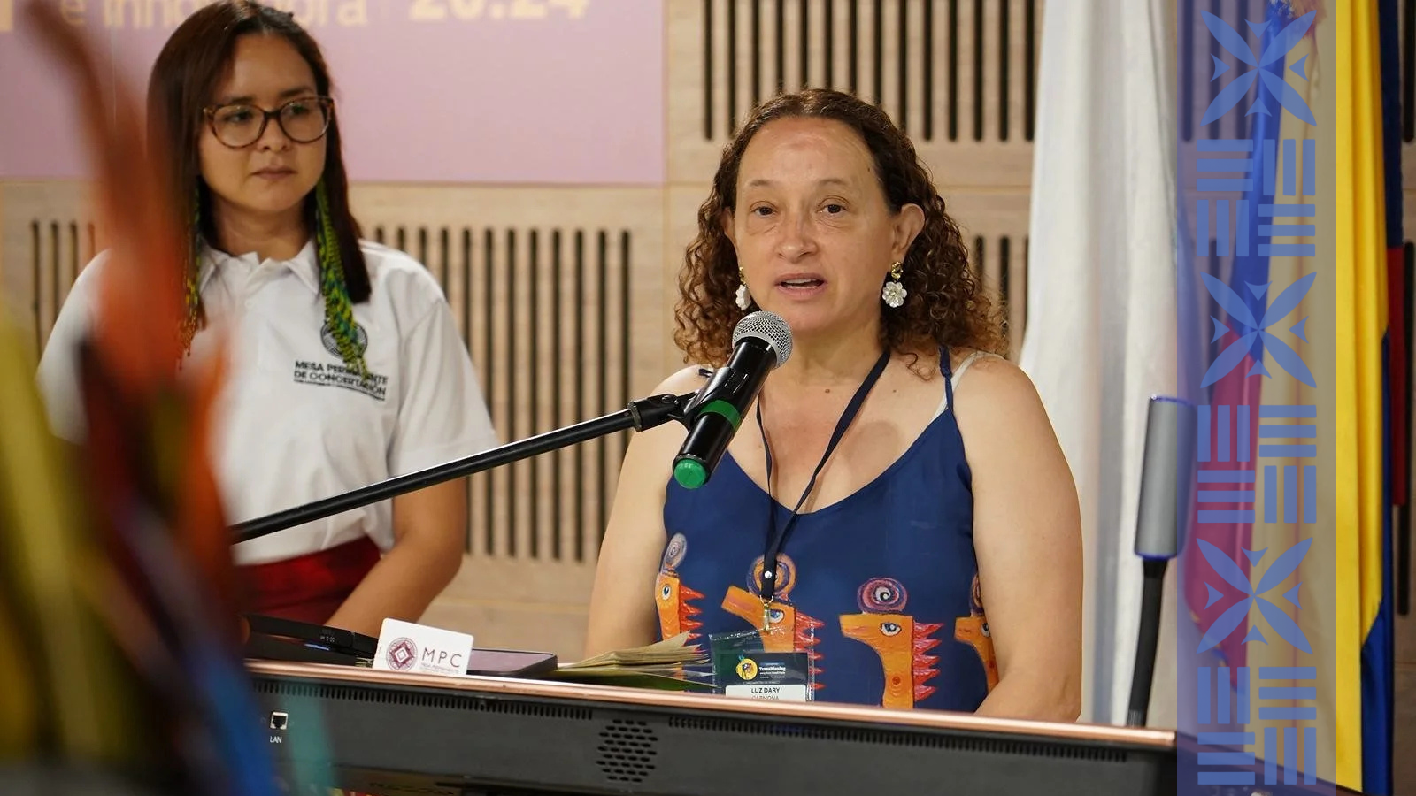 A woman with curly hair, wearing a blue dress with animal designs, speaking into a microphone at a conference. A woman in glasses and a white shirt with a name tag and an indigenous necklace is in the background.