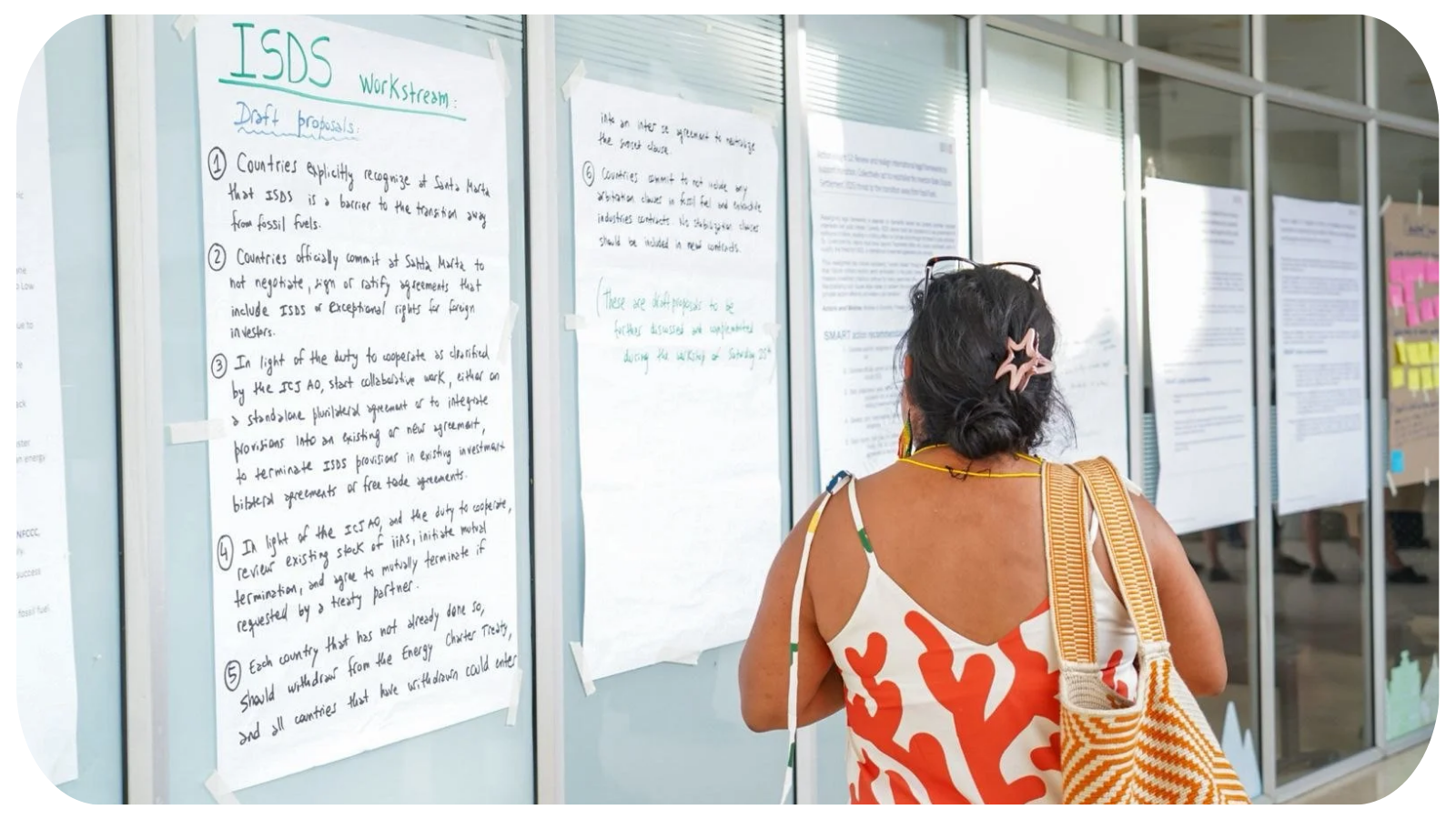 A woman in a red and white patterned dress with a yellow strap bag reads notes on a poster board at an outdoor event, with glass windows in the background.