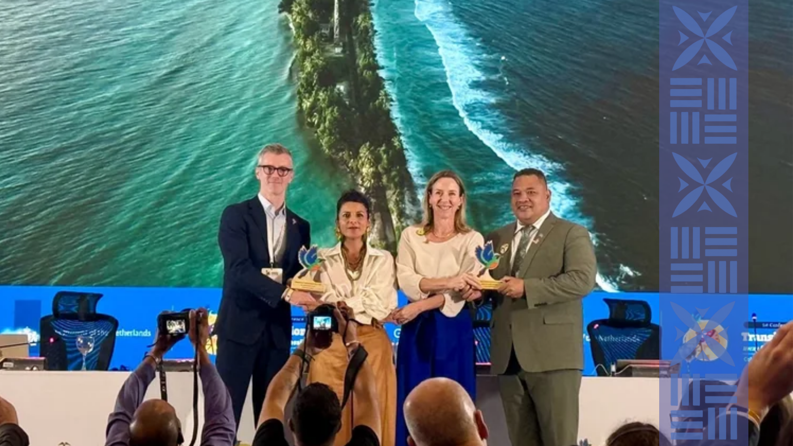 Four people wearing formal attire stand on stage, holding awards, with a large screen displaying an aerial view of land and water in the background. Audience members are taking photos.