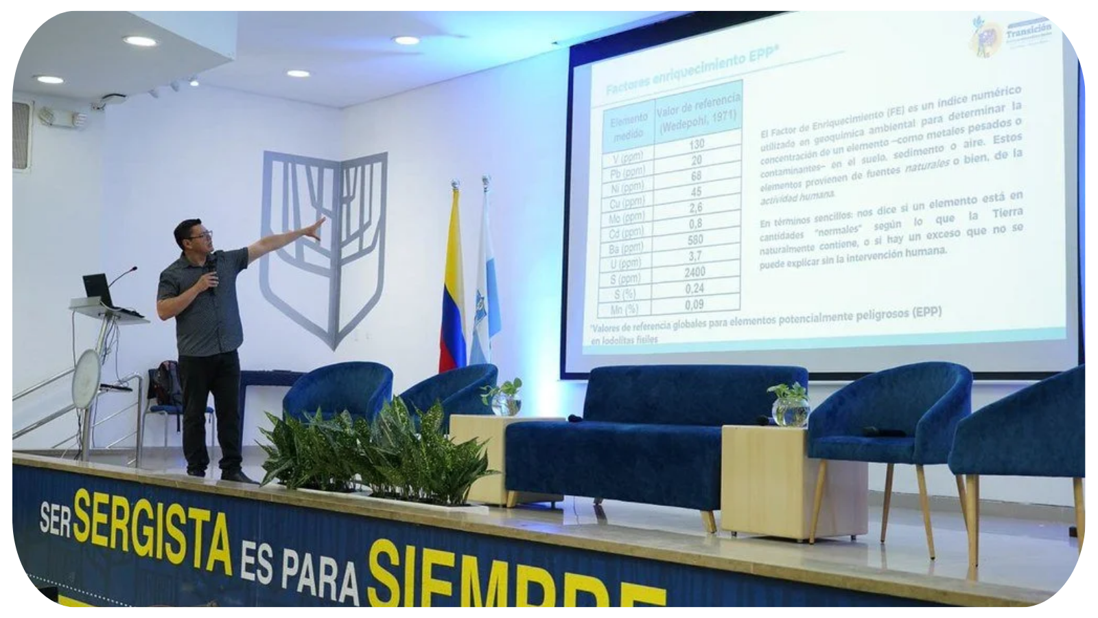 A man giving a presentation on a stage with a microphone, pointing at a large screen displaying a table of data and text. The stage has a banner with the words 'serSergista es para siempre'. There are flags of Colombia and another flag behind him, and chairs with plants in front of them.