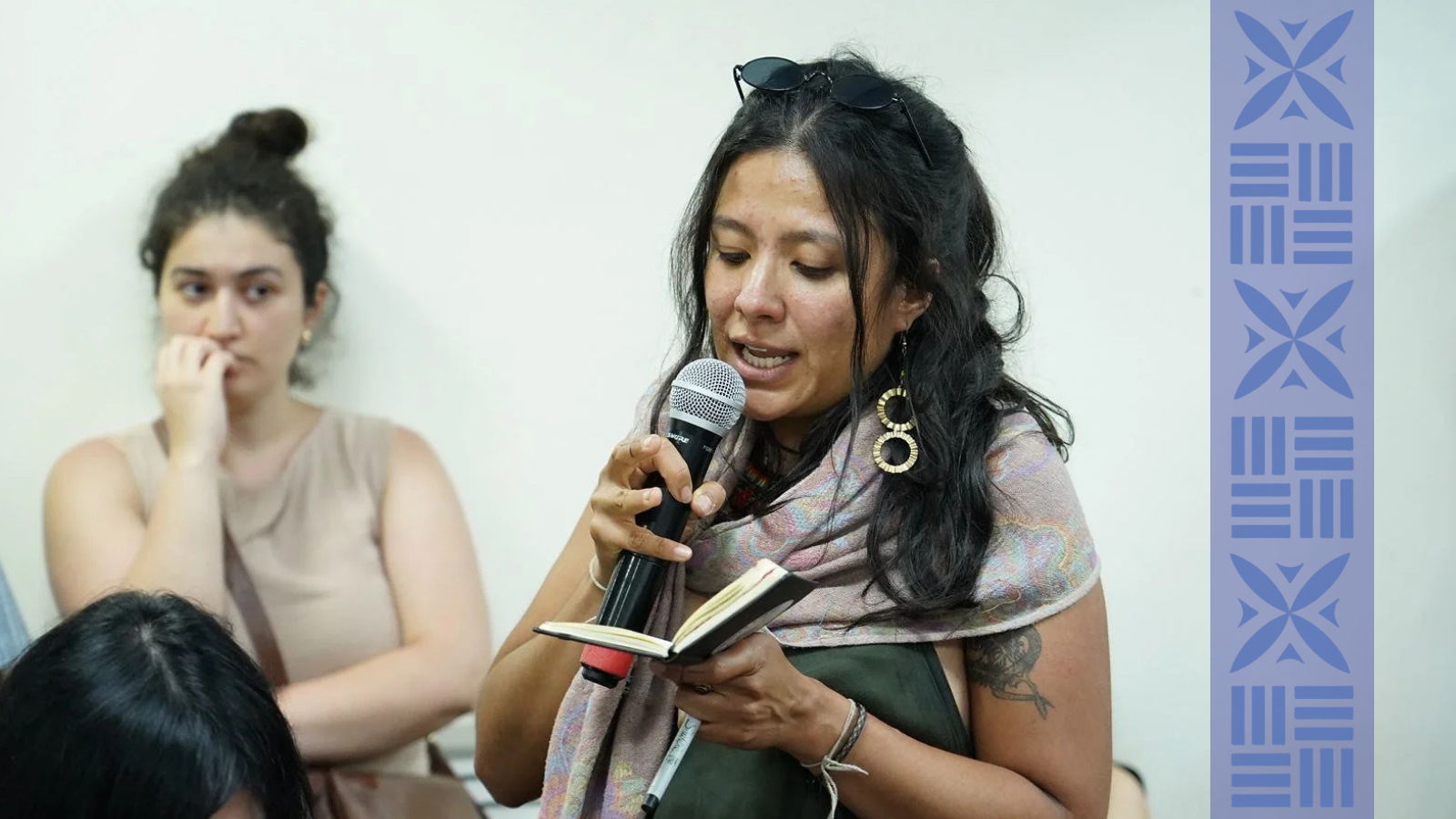 A woman with dark wavy hair, wearing earrings and a scarf, is speaking into a microphone while holding a small notebook. Behind her, another woman with dark curly hair in a bun is listening with her hand near her mouth.