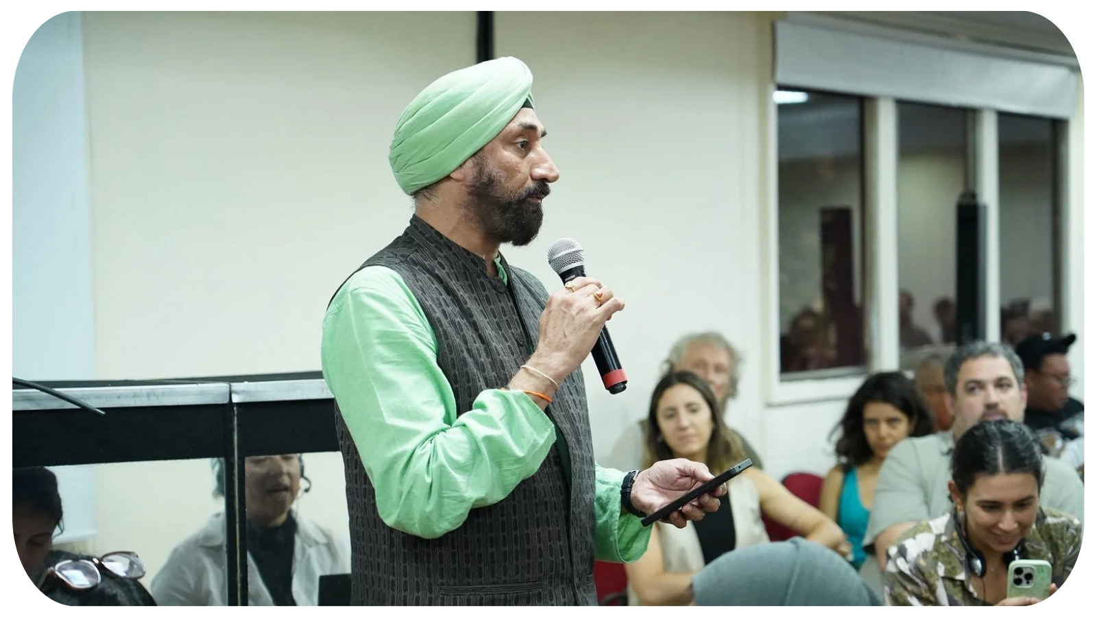Man wearing a green turban and traditional Indian attire speaking into a microphone at a conference room, with audience members listening and taking notes.