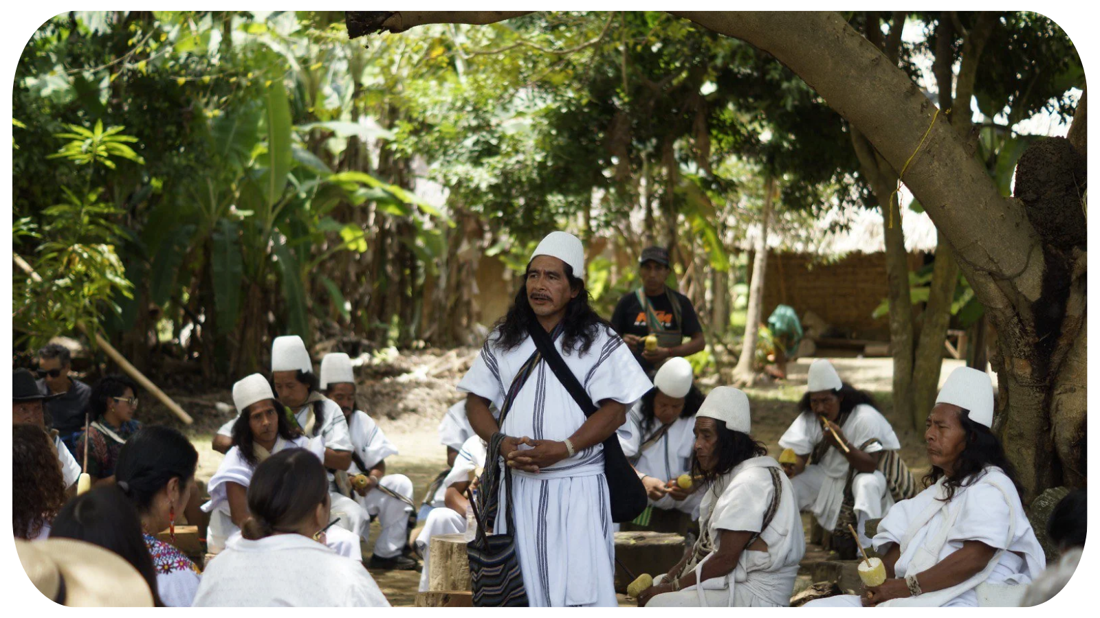 A group of indigenous men and women dressed in traditional white clothing and hats are gathered outdoors under trees. They are seated and appear to be participating in a cultural event, with some holding musical instruments and coconuts. One man in the center stands with a serious expression, possibly leading or performing.