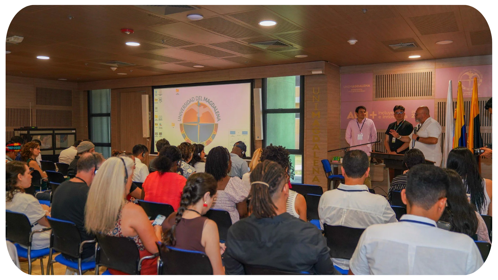 A diverse group of people attending a presentation or seminar in a conference room with a university logo on a large screen and walls, with four speakers at the front.