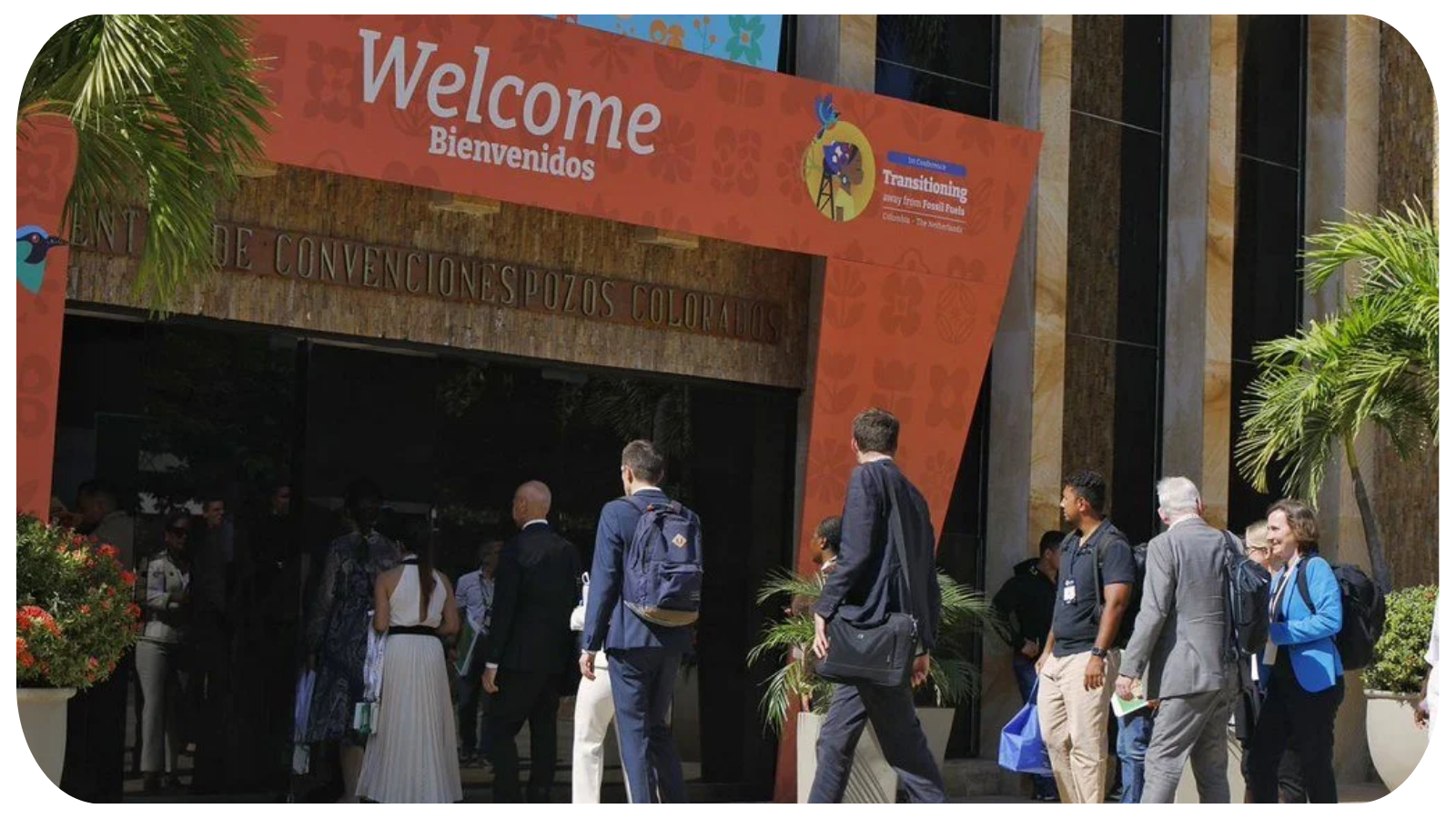 People entering a conference under a large orange banner that reads 'Welcome' and 'Bienvenidos' at the entrance of a convention center, with palm trees and decorative plants outside.