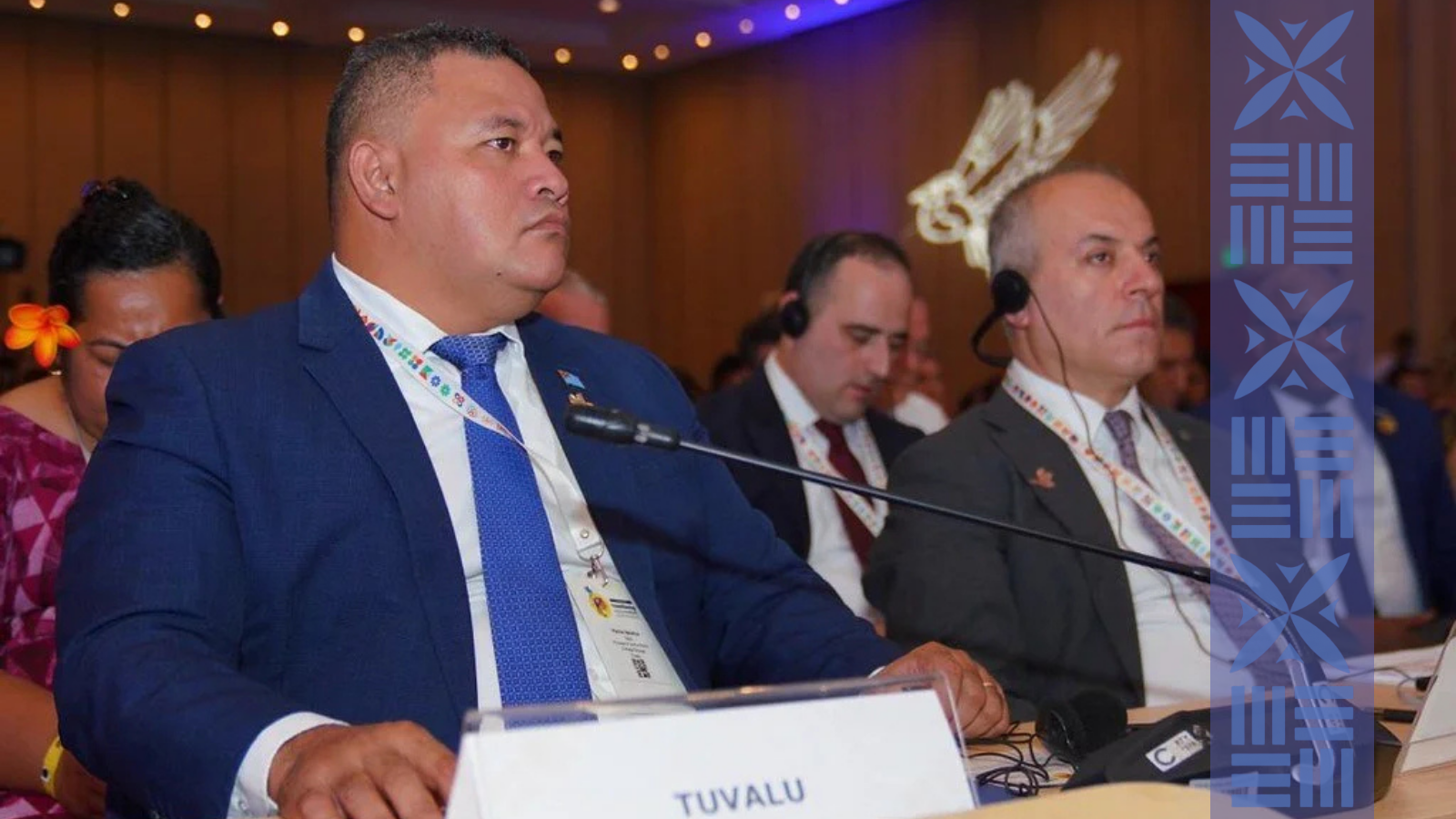 Two men sitting at a conference table during an international event, wearing formal suits with traditional necklaces, one with a traditional headpiece, and listening attentively.