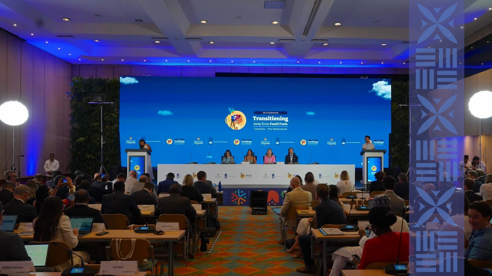 A conference room with a large stage and a blue screen displaying the title 'Transitioning away from Fossil Fuels' in Colombia, the Netherlands. Several people are seated at a panel on the stage, with a speaker at a podium on the left and right. The audience, seated at tables, is engaged, with some using laptops and taking notes. The room has bright lighting, and there are large white round lights on either side of the stage.