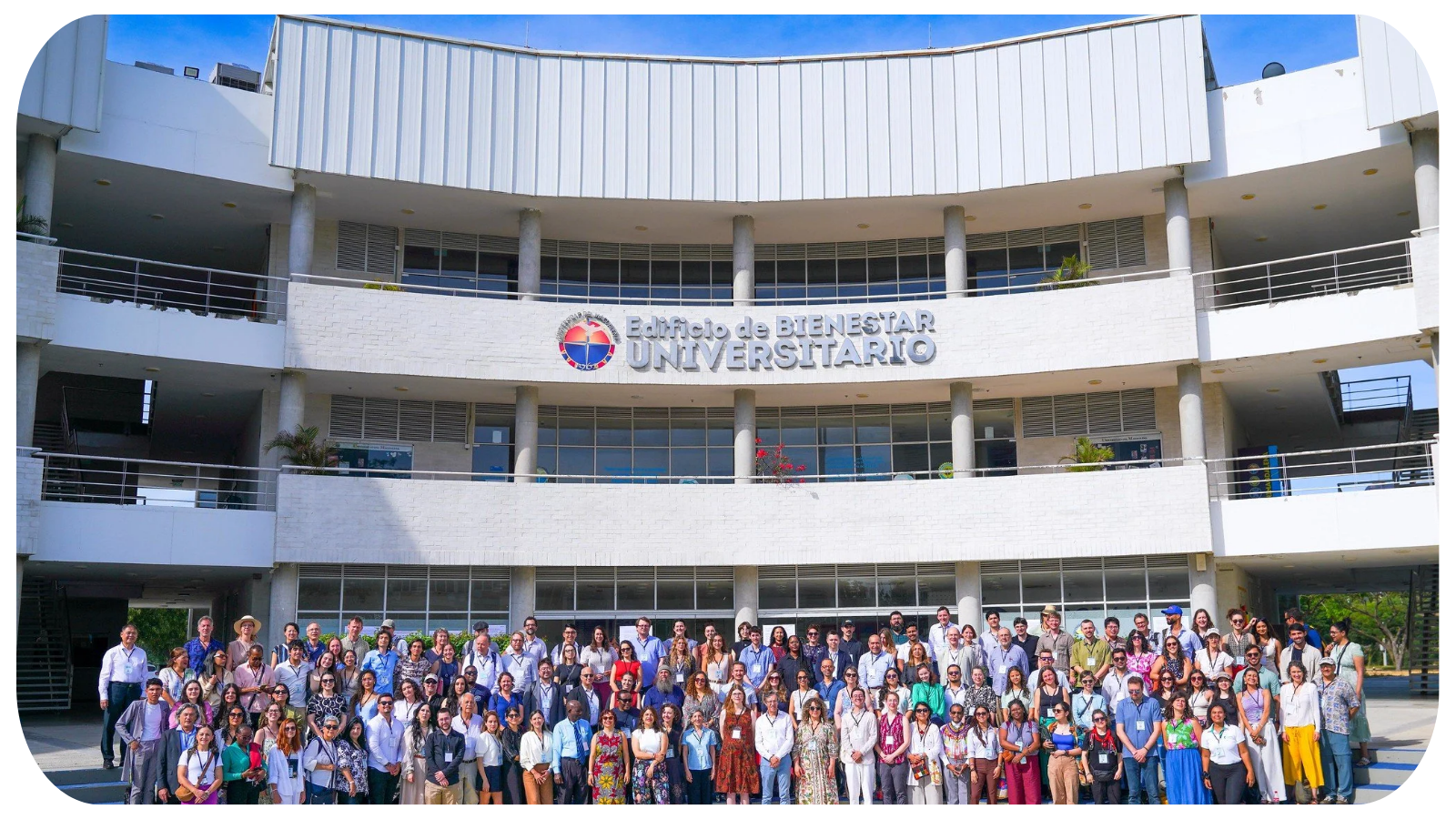 A large group of people standing in front of a modern, multi-story university building with a sign that reads 'Edificio de Bienestar Universitario' and a colorful logo above it.
