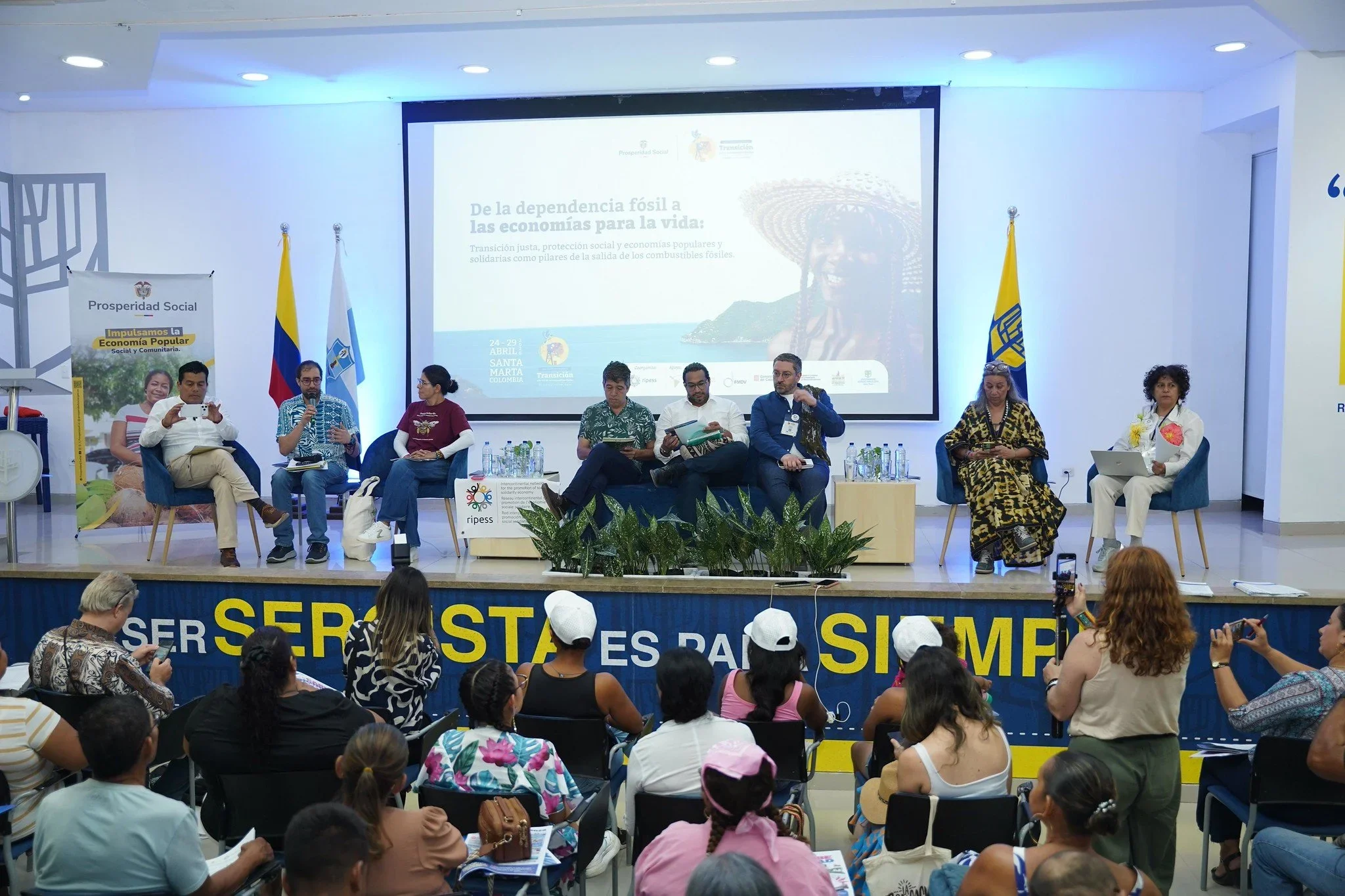 A panel discussion on stage with eight speakers, including men and women, in front of a large presentation screen. Audience members are seated and standing, some taking photos or videos, in a conference room decorated with flags and banners promoting social prosperity and sustainable development.
