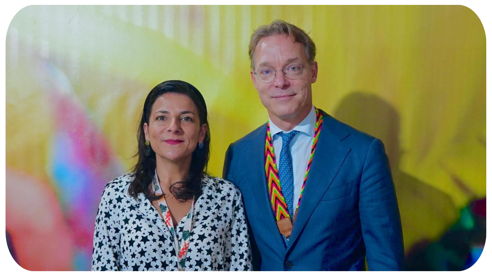 A woman and a man standing side by side, smiling, in front of a yellow background at a formal event.