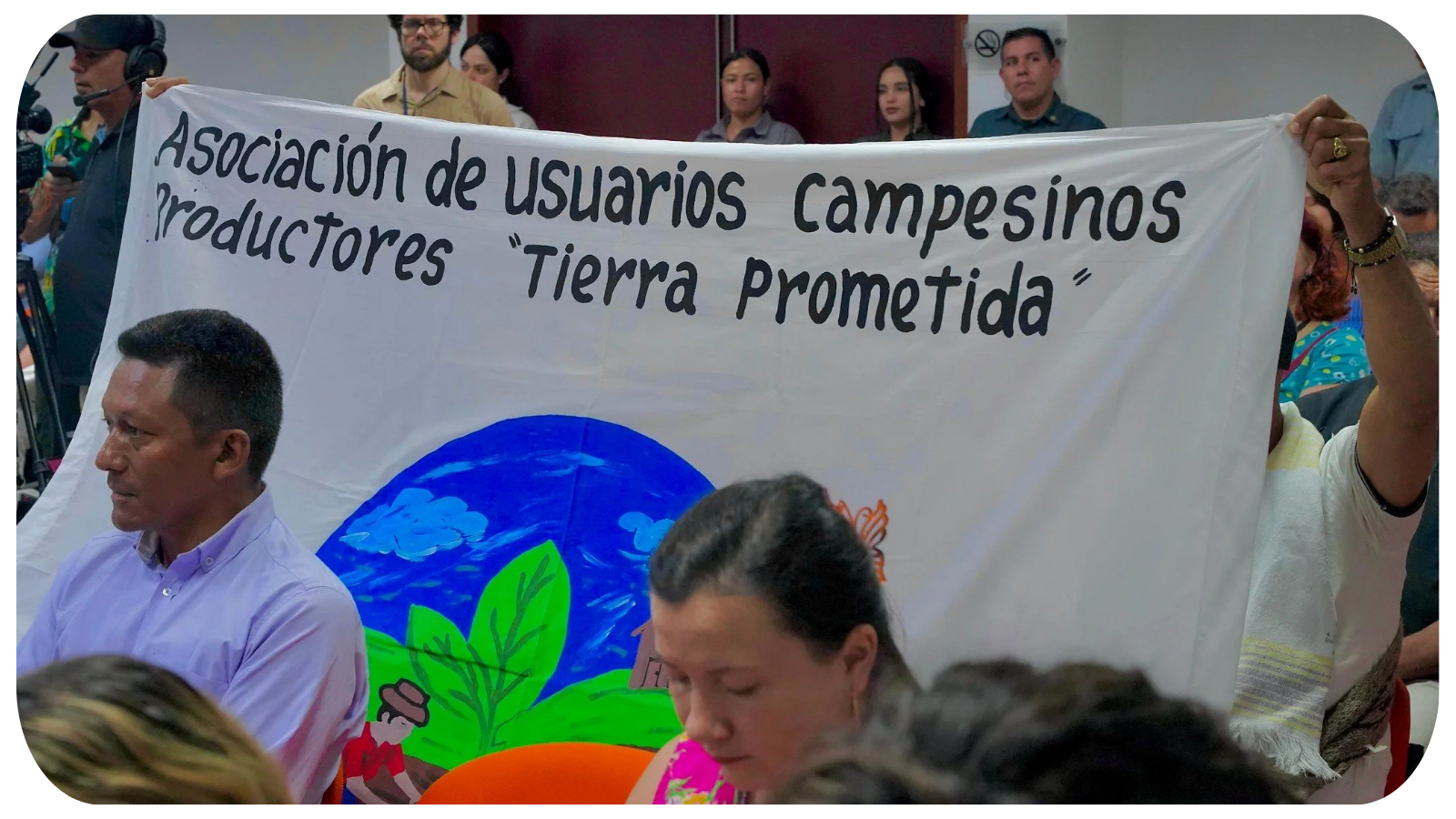 People gathered at a meeting holding a large banner that reads 'Asociación de Usuarios Campesinos Productores Tierra Prometida' with a colorful drawing of a landscape, including a farmer, plants, and a blue sky.