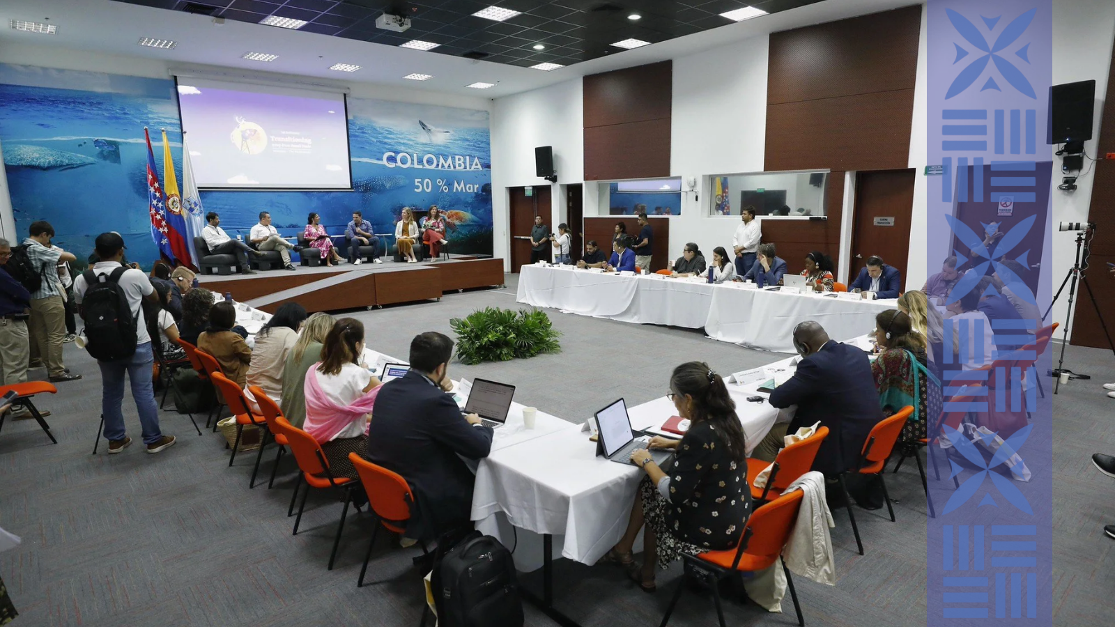 A conference or panel discussion taking place in a large room. Several people are seated at a long table facing the audience, with multiple flags behind them. Attendees are sitting and working on laptops, some are standing or filming, and a large screen displays a presentation with ocean-themed visuals and the word 'COLOMBIA' in the background.