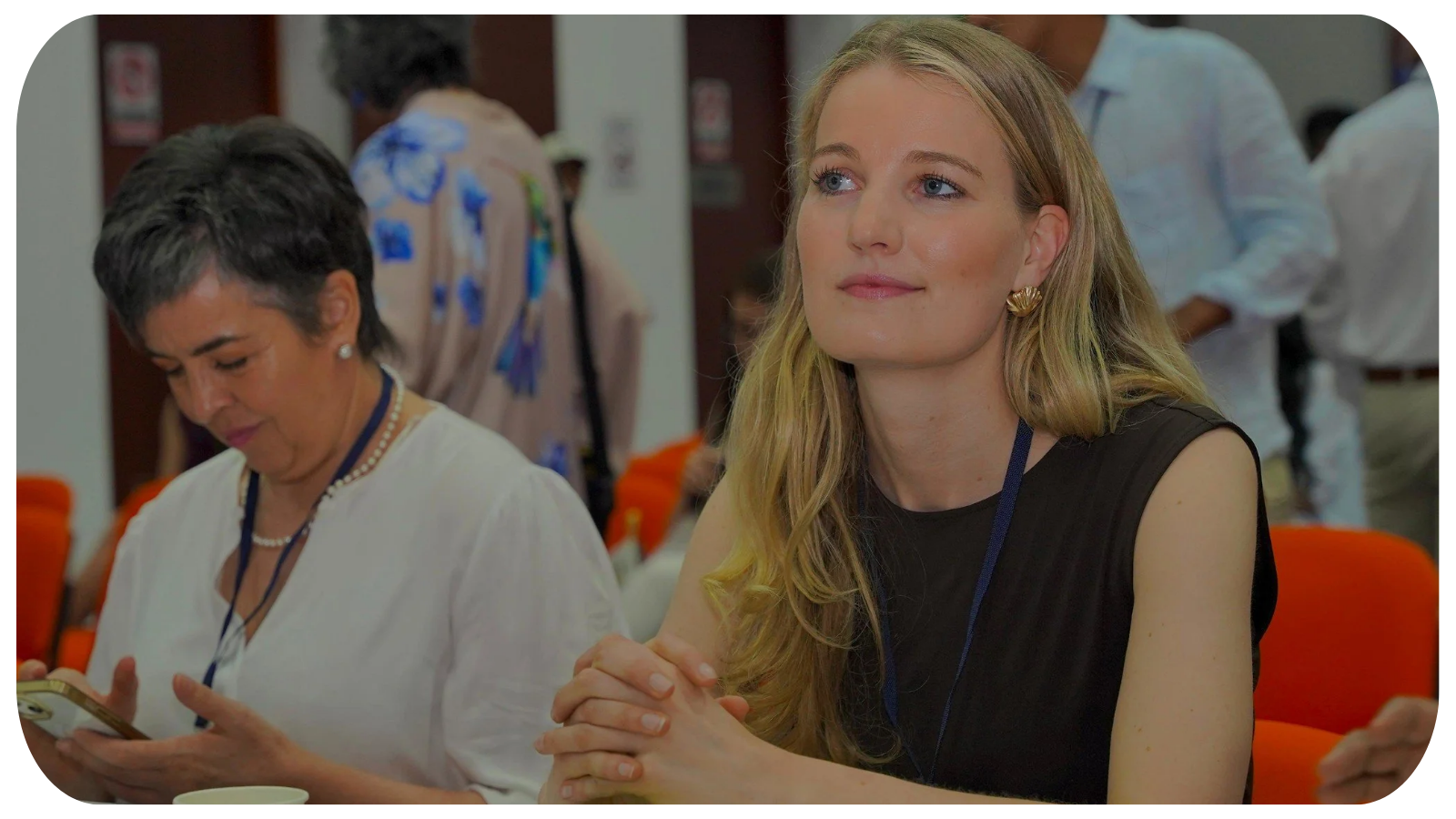A woman with blonde hair and a black sleeveless top sits at a table with her hands clasped, looking thoughtfully. Beside her, a woman with short gray hair, wearing a white blouse and pearl jewelry, is looking at her phone. The background shows other people engaging in conversations in a conference room setting.