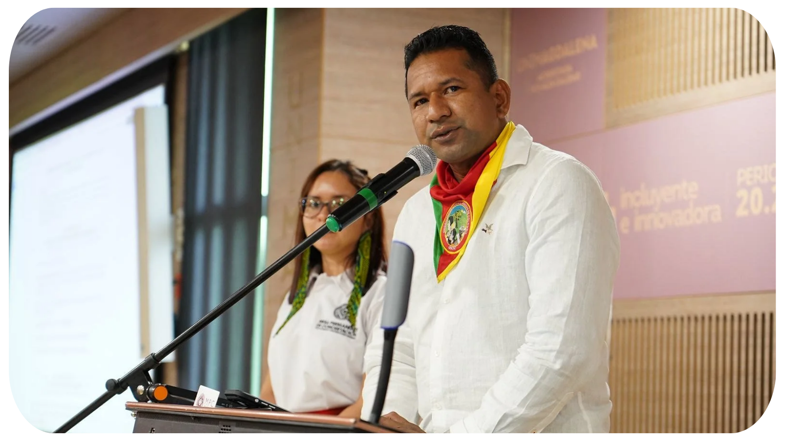 A man speaking at a podium with a microphone, wearing a white shirt and a colorful scarf, with a woman standing behind him. They are at a conference or meeting, with a blurred presentation screen and a background banner.