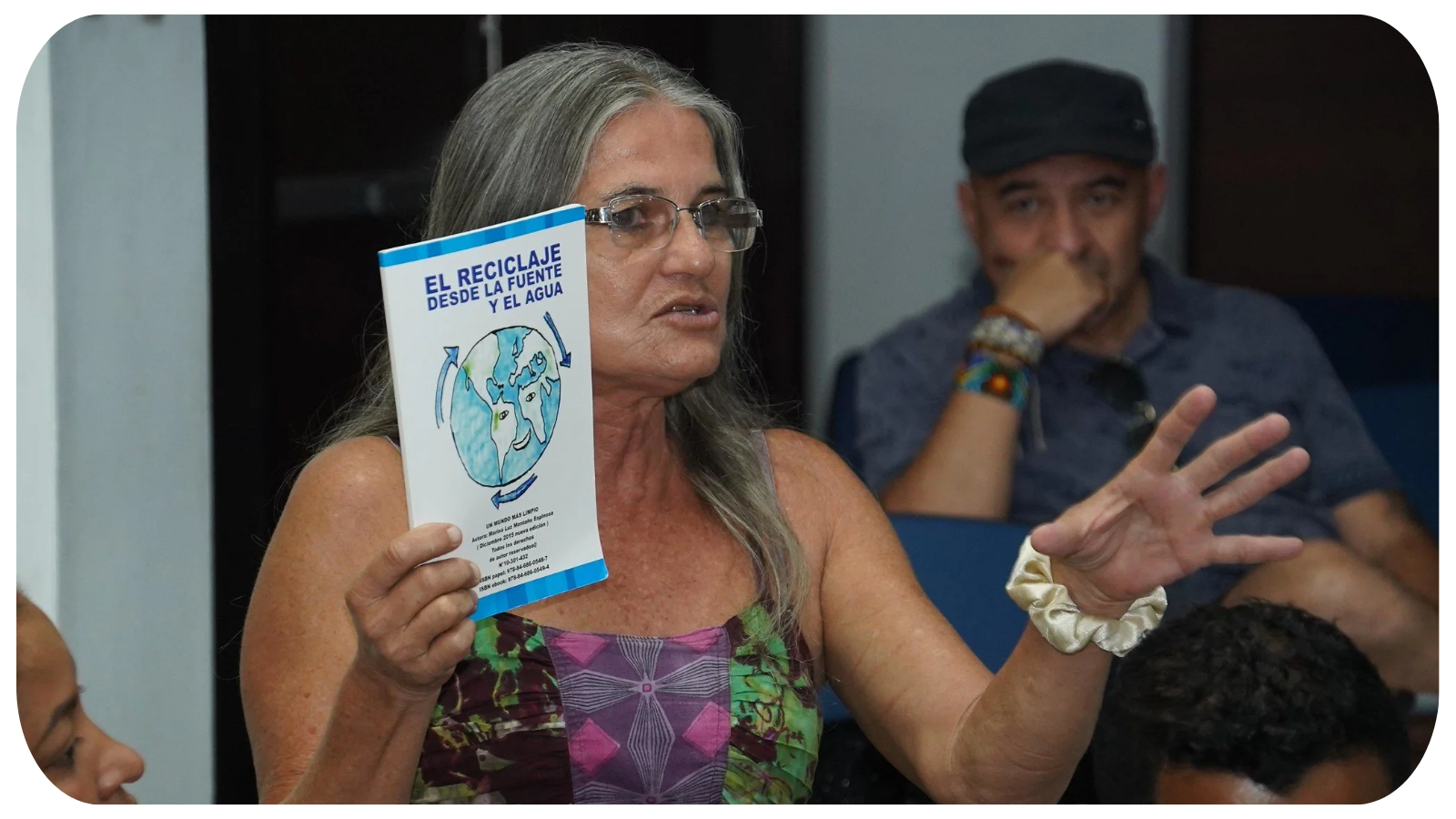 An elderly woman with long gray hair and glasses holding a pamphlet about recycling, with a man in the background resting his chin on his hand.