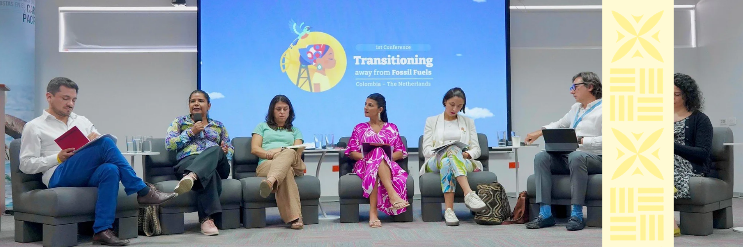 Six people sitting on a row of chairs in a conference room, participating in a panel discussion. There are five women and one man. A large screen behind them displays the conference title: "Transitioning away from Fossil Fuels, Colombia - The Netherlands." The room has a modern appearance with a yellow patterned side panel and glasses of water on a table behind the panel members.
