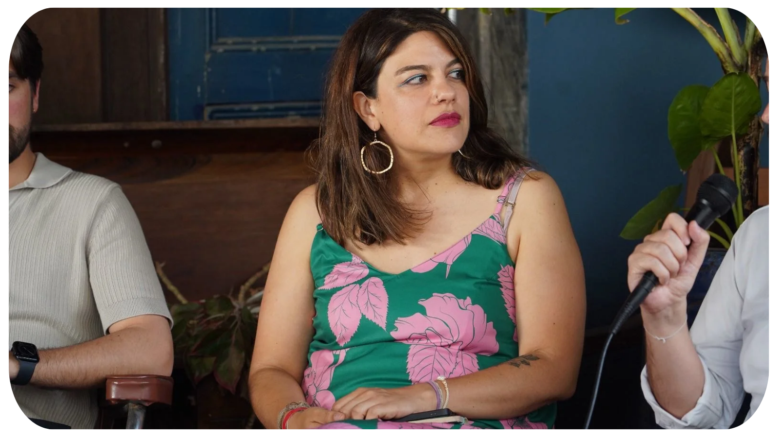 A woman with shoulder-length brown hair wearing a green and pink floral dress, large hoop earrings, and colorful bracelets, holding a smartphone and listening attentively during a discussion, with a person holding a microphone nearby.
