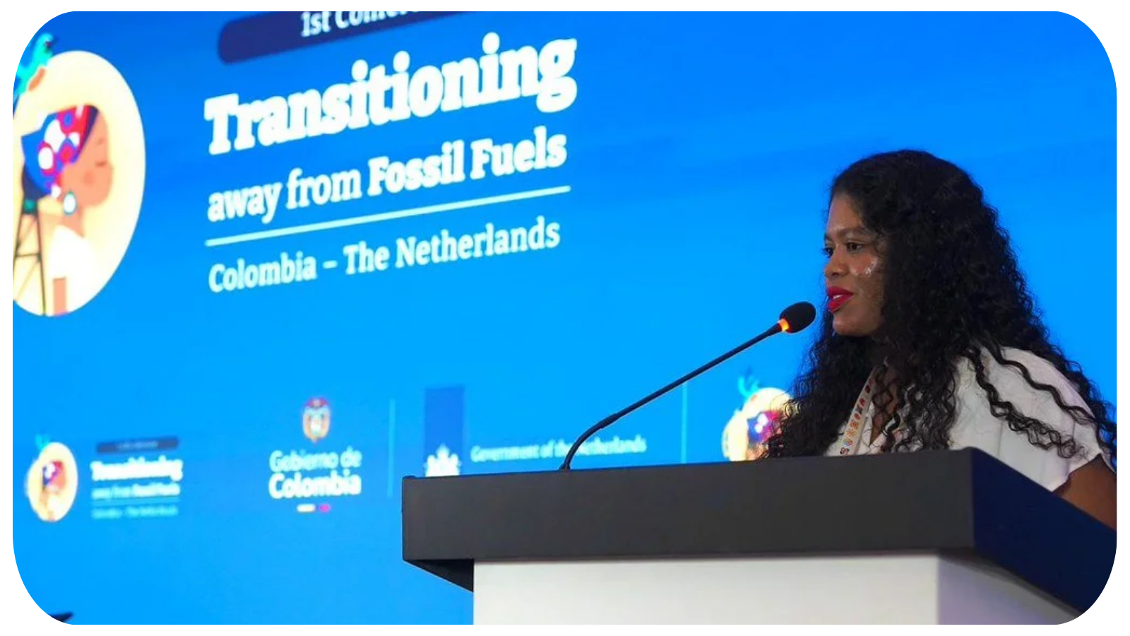 A woman with dark curly hair and red lipstick giving a speech at a conference with a large blue presentation screen behind her. The screen displays a title about transitioning away from fossil fuels, mentioning Colombia and the Netherlands.