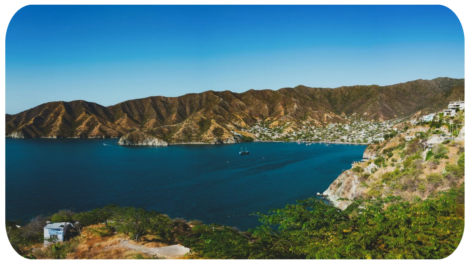 A coastal bay surrounded by hills with residential buildings, boats on the water, and greenery in the foreground under a clear blue sky.