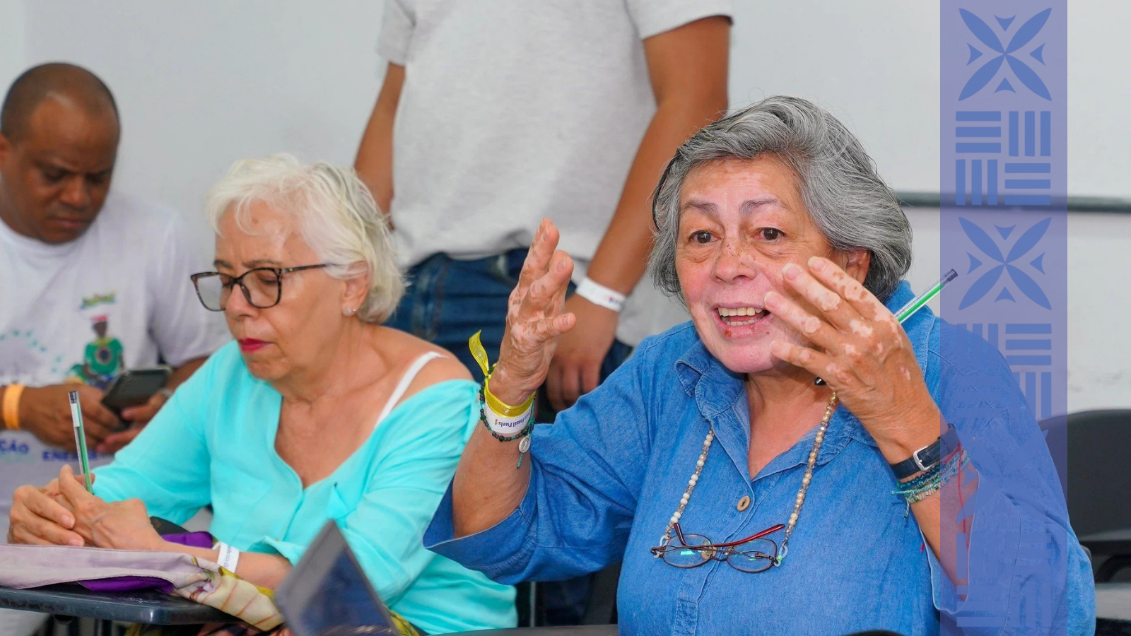 A group of elderly people sitting at a table in a classroom, engaging in a discussion. The woman on the right, with gray hair and glasses hanging from her shirt, is speaking animatedly, gesturing with her hand. The woman next to her is writing, while a man in the background is looking at his phone. A person in a gray shirt is standing behind them.