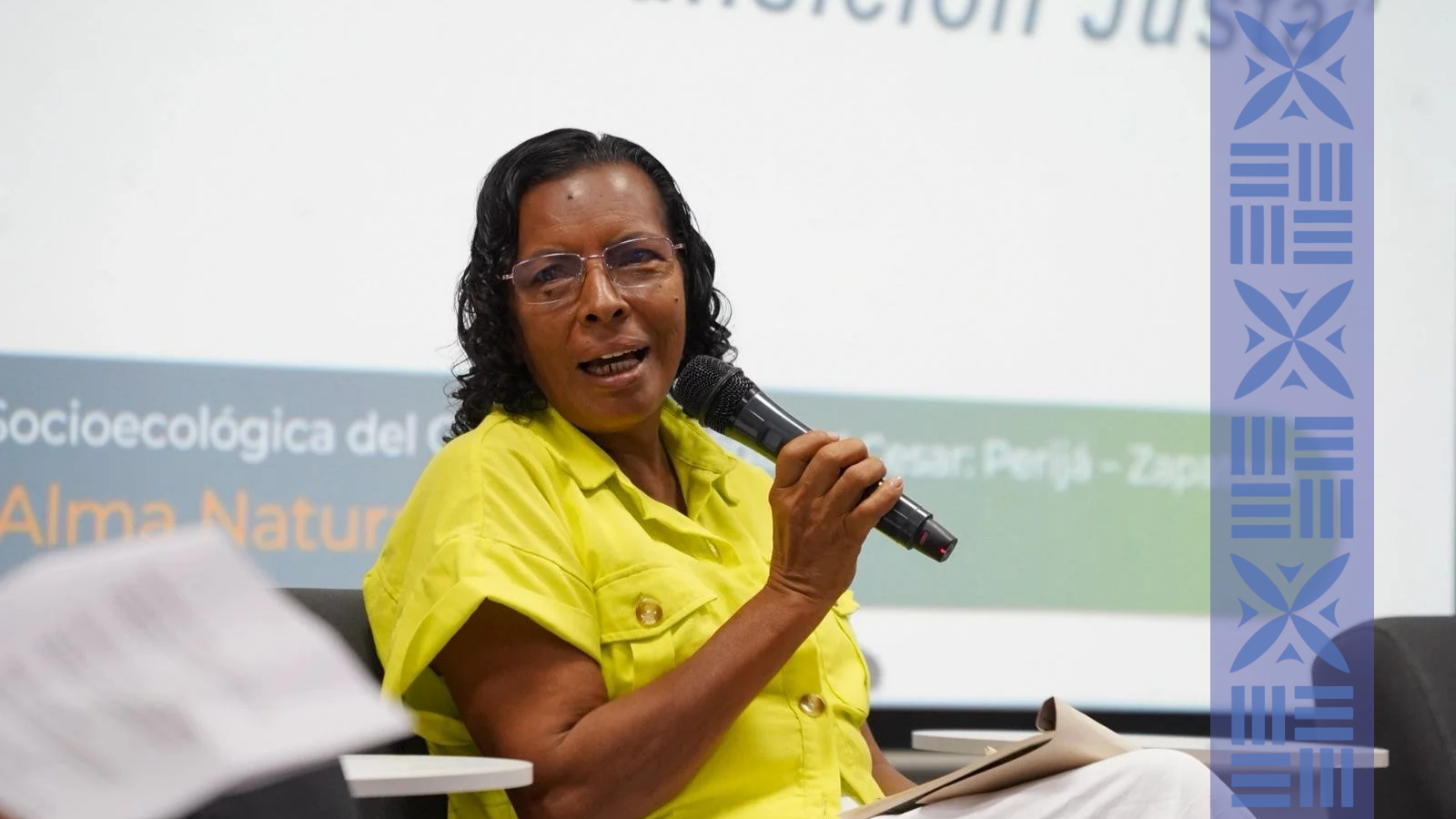 A woman with curly black hair, wearing glasses and a yellow shirt, is holding a microphone and speaking at a conference or seminar. There is a presentation screen behind her with text in Spanish.