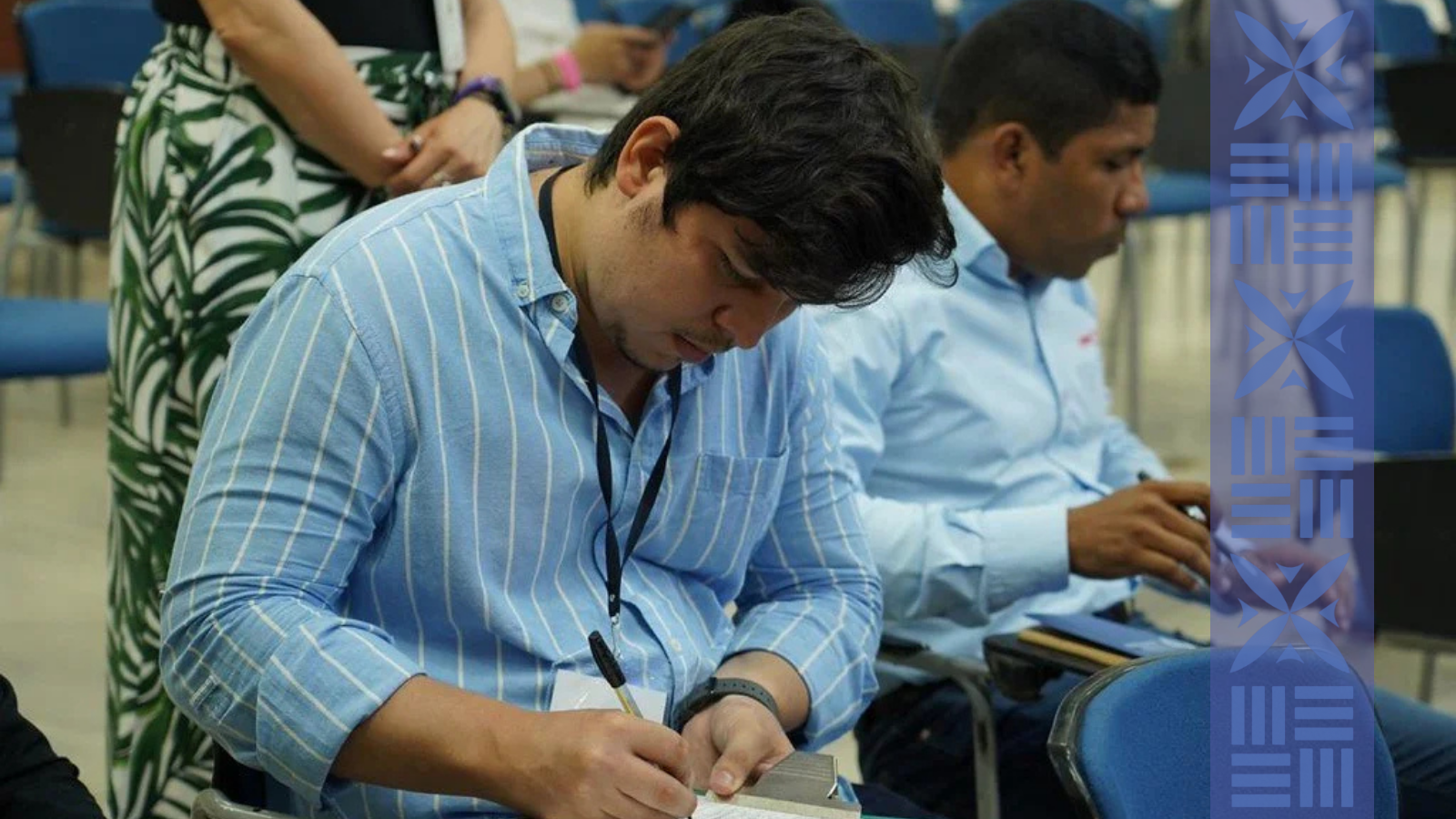 A man with dark hair writing in a notebook at a conference or seminar, with another man with short dark hair sitting next to him, both wearing button-up shirts, and a woman standing behind them.
