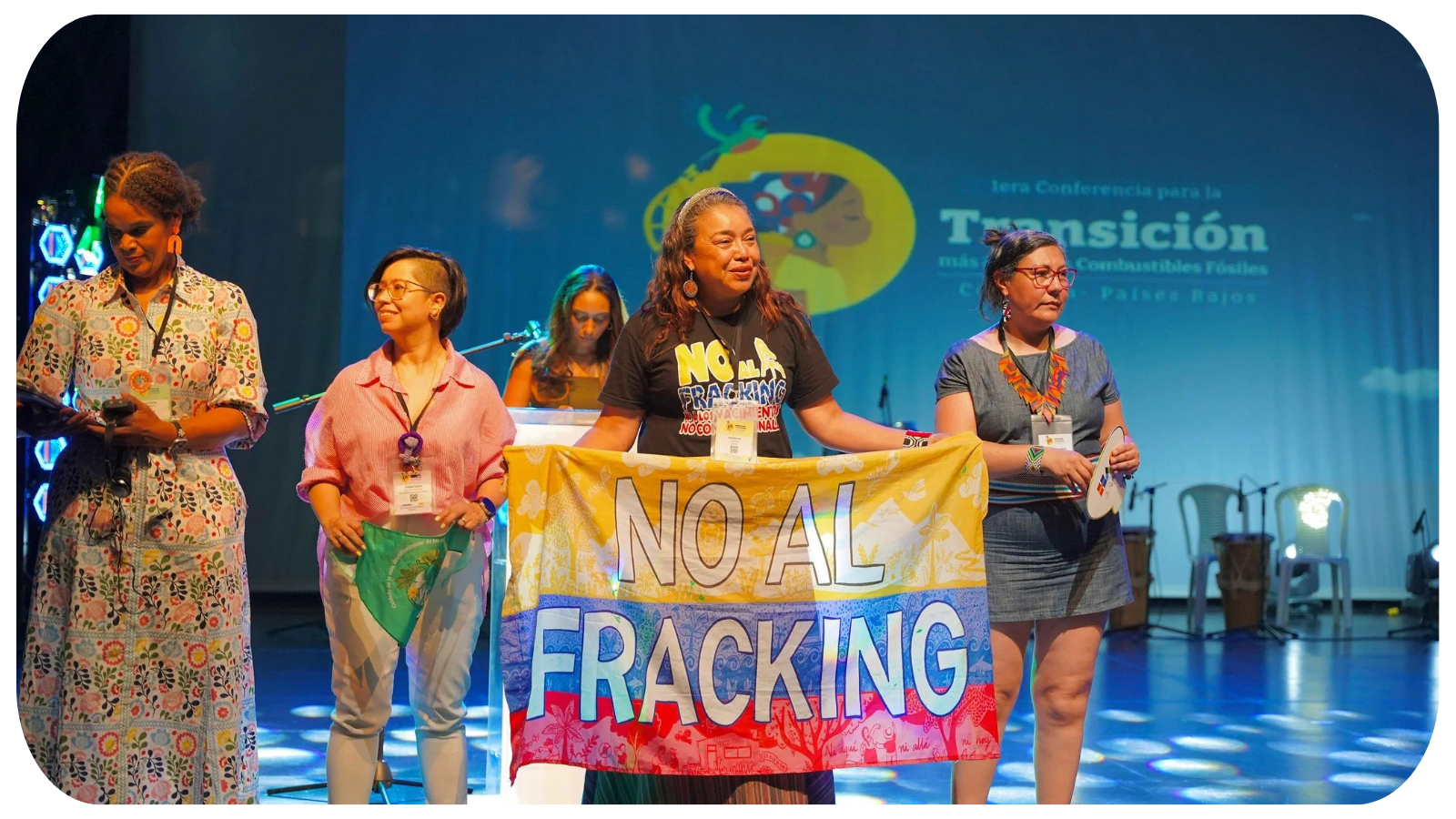 Women standing on stage holding a colorful banner that reads "NO AL FRACKING," during a conference about fossil fuels and transformation.