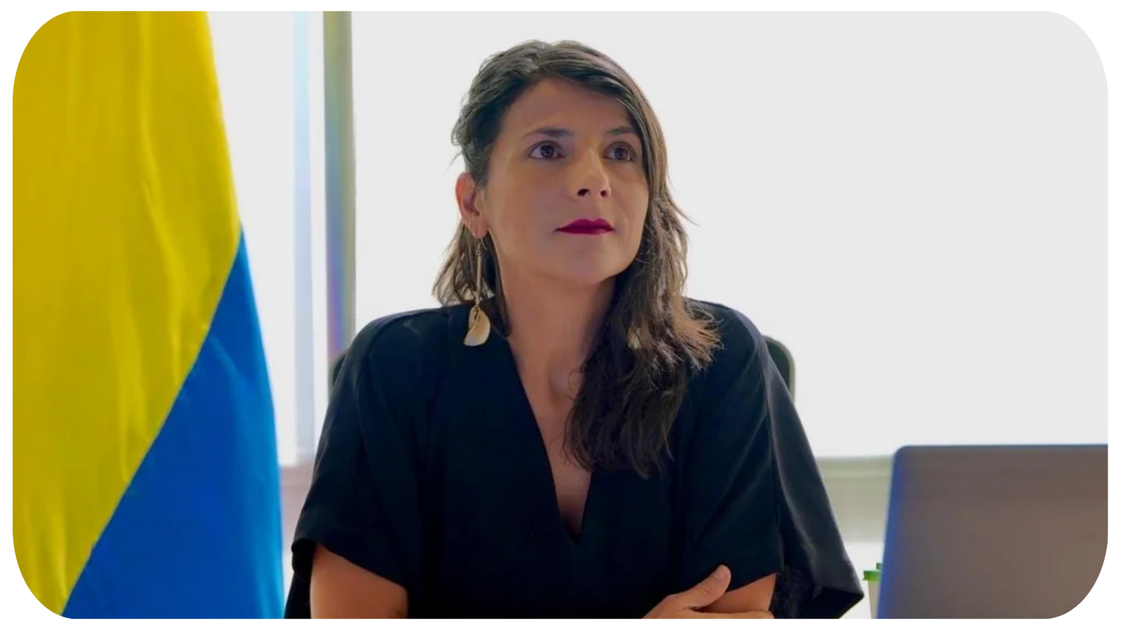 A woman with dark brown hair, wearing a black top, sitting at a desk in an office. Part of a blue and yellow flag is visible on her left. There is a laptop in front of her. She is looking slightly to the right with a serious expression.